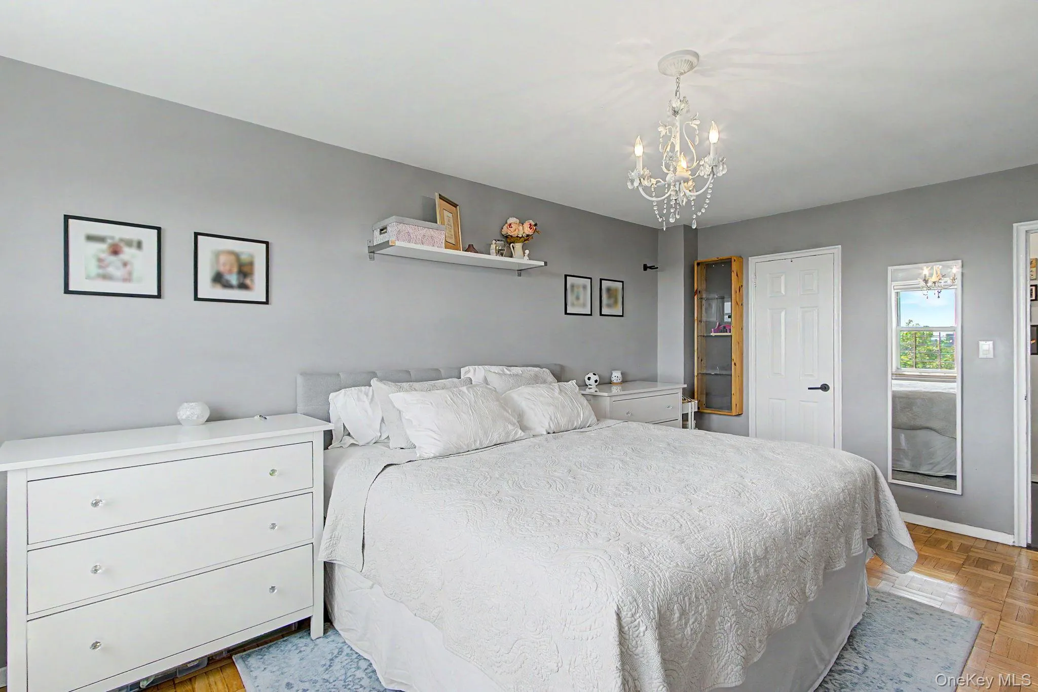 Bedroom featuring a crystal chandelier, light gray walls, wood-finish flooring, and a built-in wall cabinet with glass doors Bedroom featuring a crystal chandelier, light gray walls, wood-finish flooring, and a built-in wall cabinet with glass doors