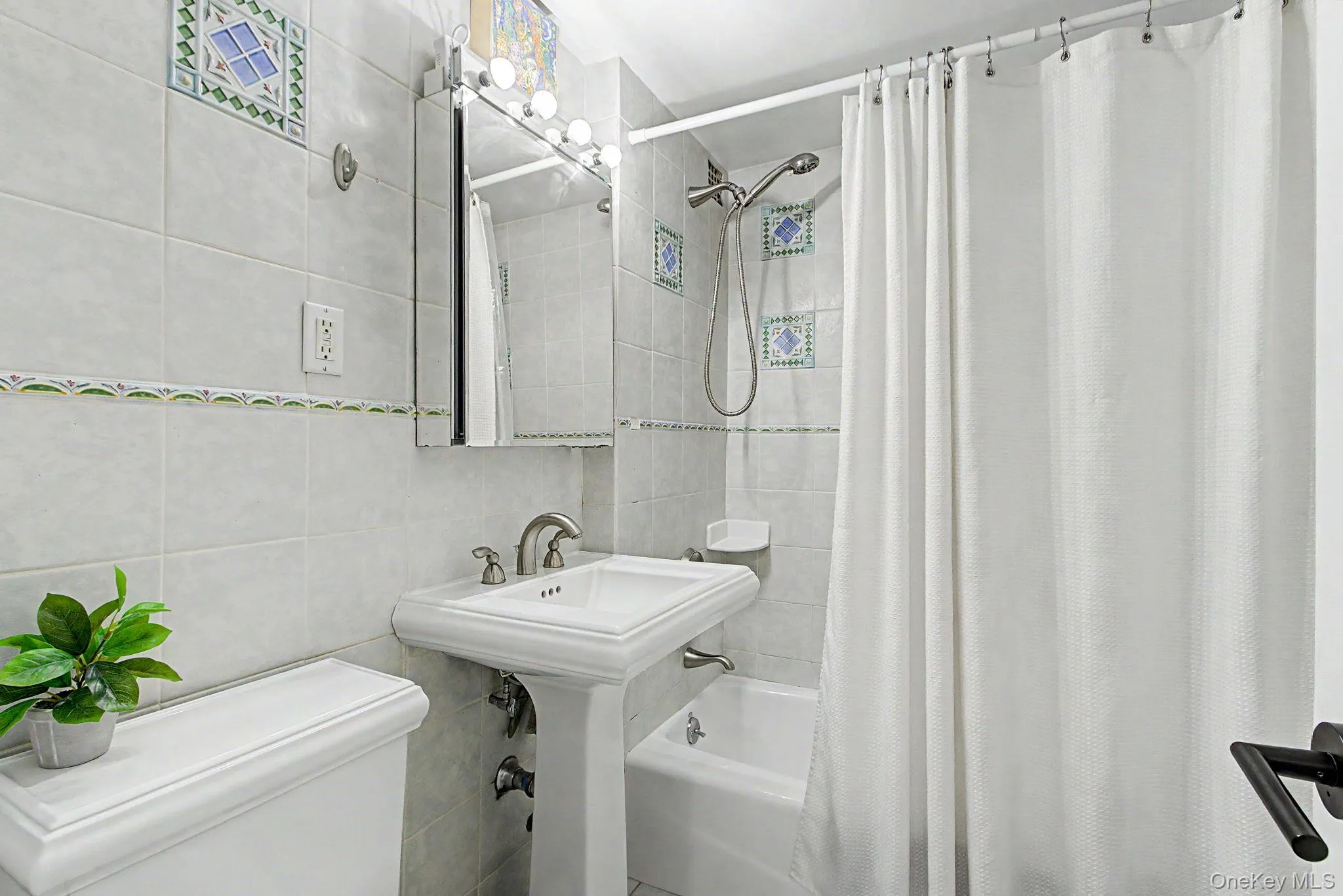 Bathroom featuring floor-to-ceiling light-toned tile, a pedestal sink with brushed nickel fixtures, a framed mirrored medicine cabinet, and a tiled shower with a handheld showerhead Bathroom featuring floor-to-ceiling light-toned tile, a pedestal sink with brushed nickel fixtures, a framed mirrored medicine cabinet, and a tiled shower with a handheld showerhead