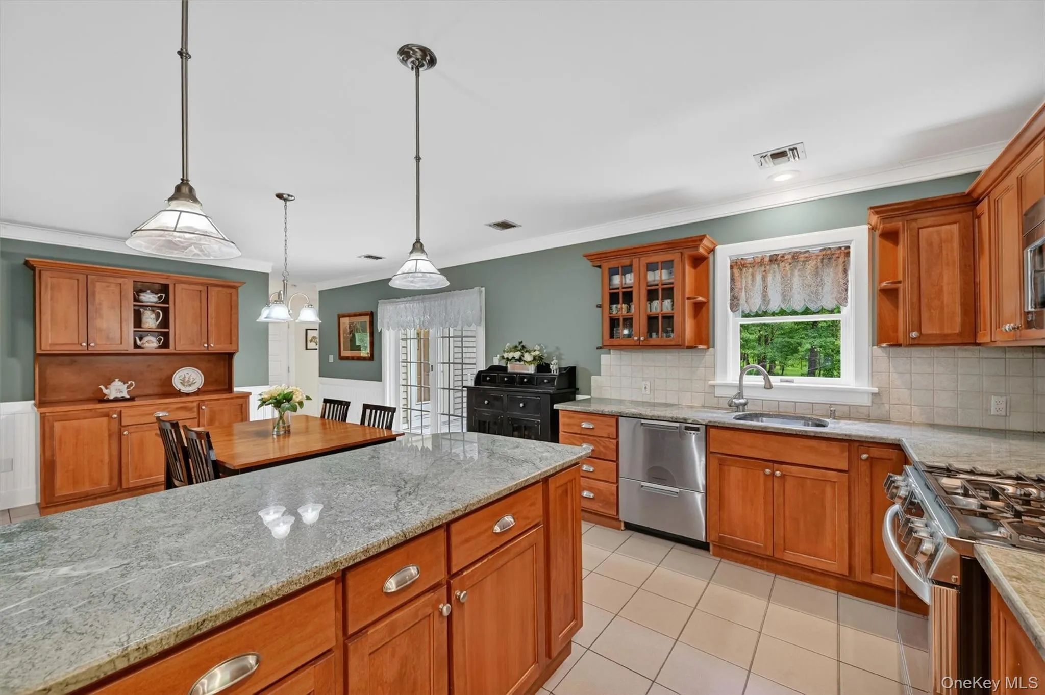 Kitchen featuring a central island with a light-toned countertop, wood cabinetry, stainless steel appliances, tile flooring, and decorative pendant lighting Kitchen featuring a central island with a light-toned countertop, wood cabinetry, stainless steel appliances, tile flooring, and decorative pendant lighting