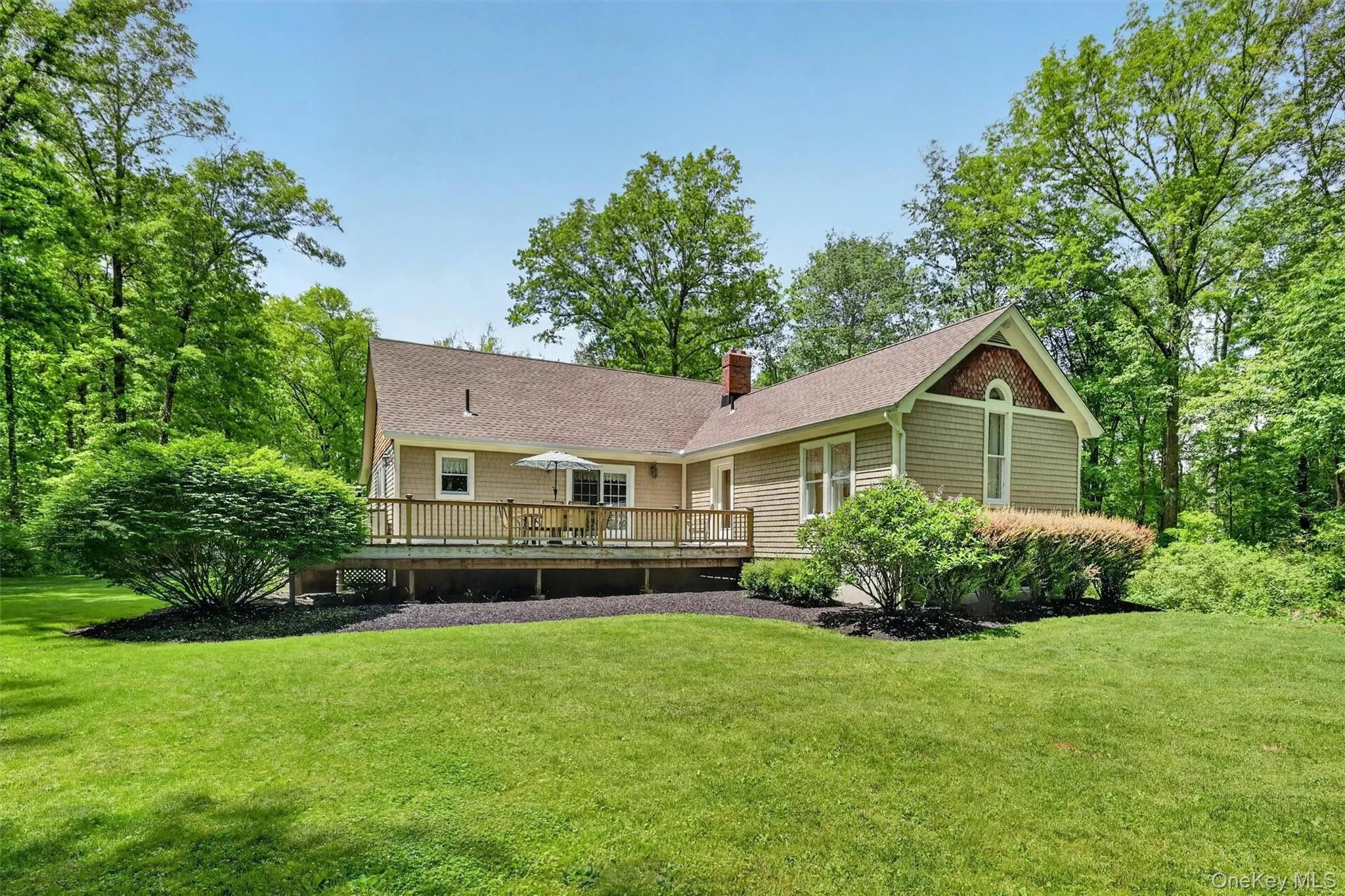Expansive green lawn surrounds a residence featuring a spacious wooden deck with railings, light-colored siding, and a prominent gable with architectural detailing Expansive green lawn surrounds a residence featuring a spacious wooden deck with railings, light-colored siding, and a prominent gable with architectural detailing