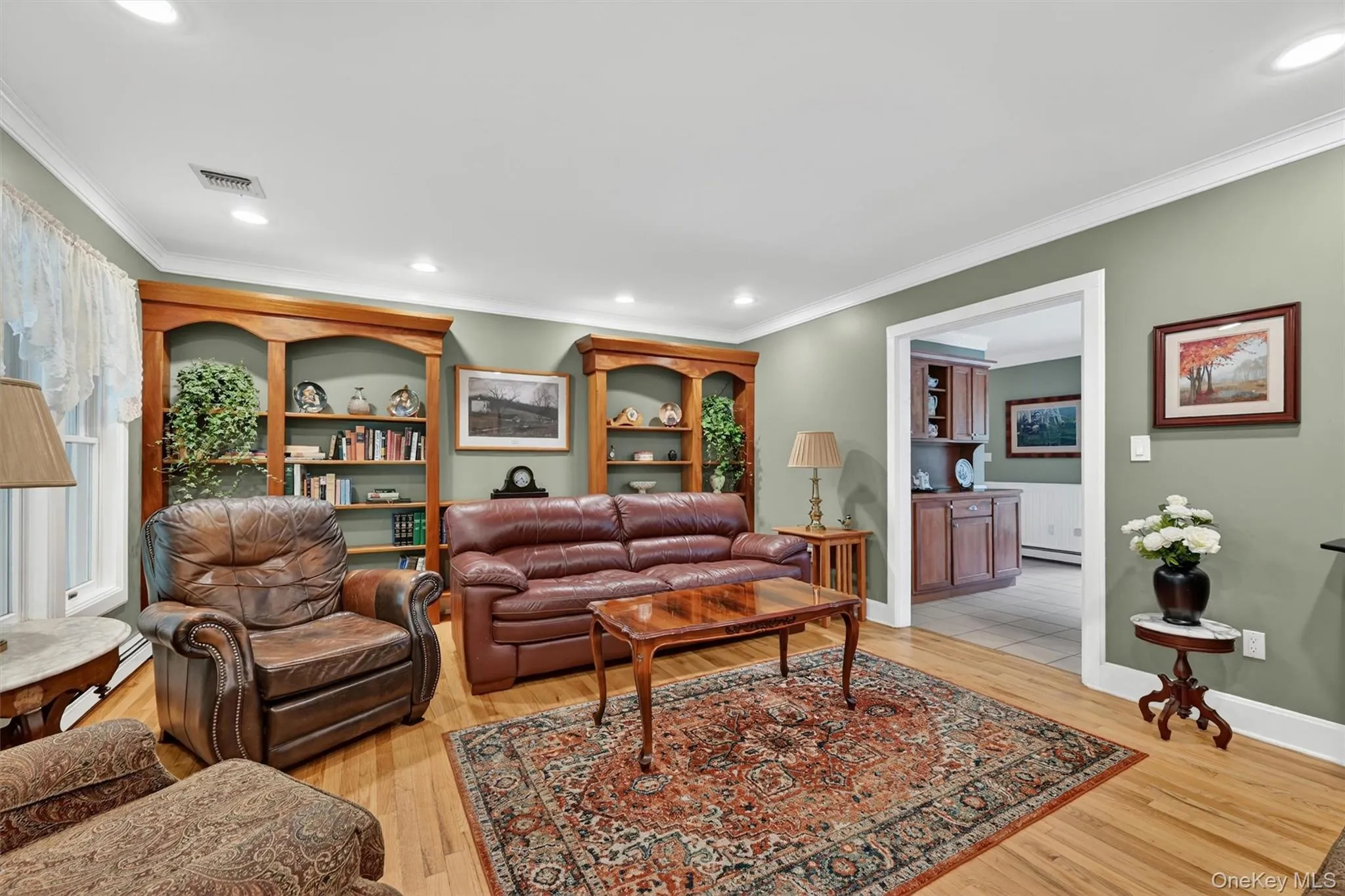 Living space featuring warm-toned wood-finish flooring, built-in display shelving, recessed lighting, crown molding, and a wide cased opening Living space featuring warm-toned wood-finish flooring, built-in display shelving, recessed lighting, crown molding, and a wide cased opening