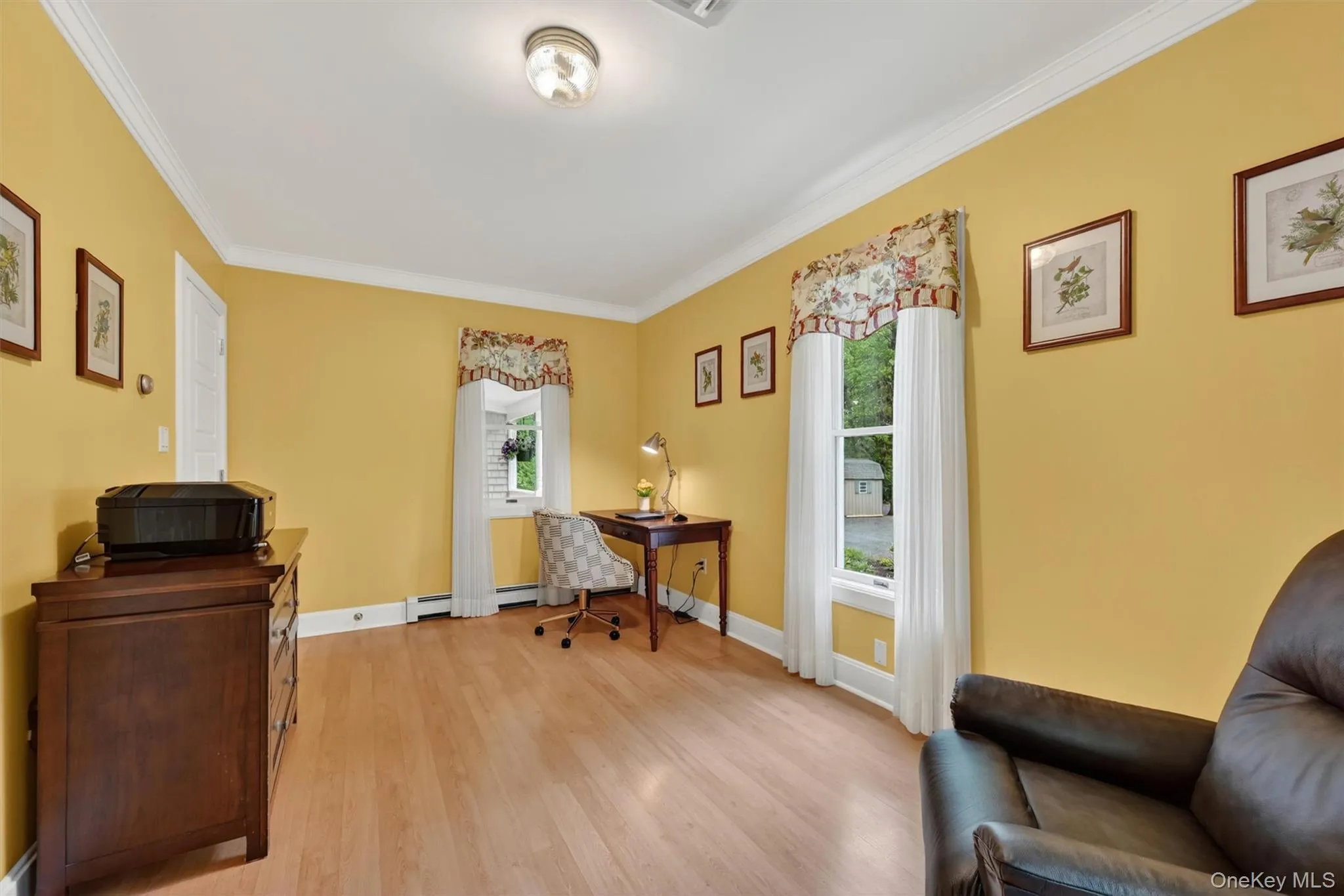 Bright room featuring wood-finish flooring, two windows with white curtains, white crown molding, and a flush-mount ceiling light fixture Bright room featuring wood-finish flooring, two windows with white curtains, white crown molding, and a flush-mount ceiling light fixture