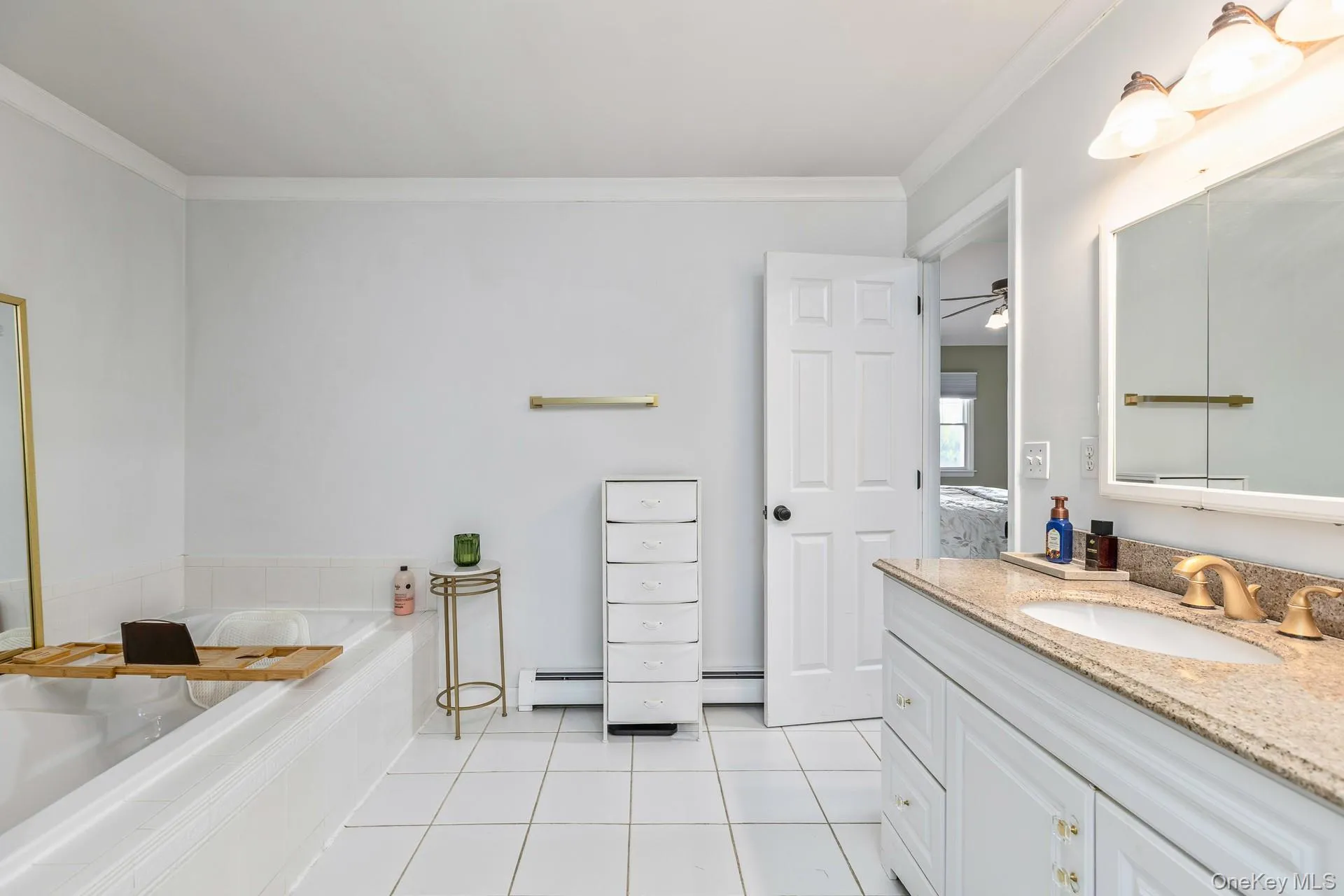 Spacious bathroom featuring a built-in soaking tub, a vanity with a light-toned stone countertop and undermount sink, and white tiled flooring Spacious bathroom featuring a built-in soaking tub, a vanity with a light-toned stone countertop and undermount sink, and white tiled flooring