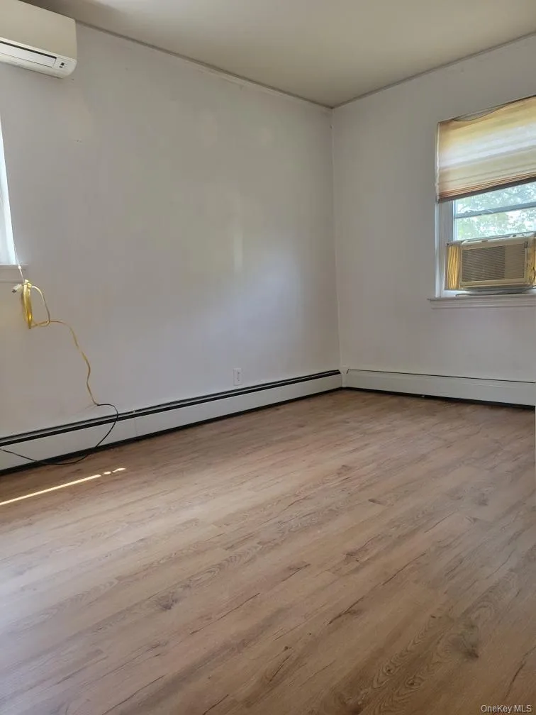 Unfurnished room featuring a wall mounted air conditioner, a baseboard radiator, wood finished floors, and cooling unit Unfurnished room featuring a wall mounted air conditioner, a baseboard radiator, wood finished floors, and cooling unit