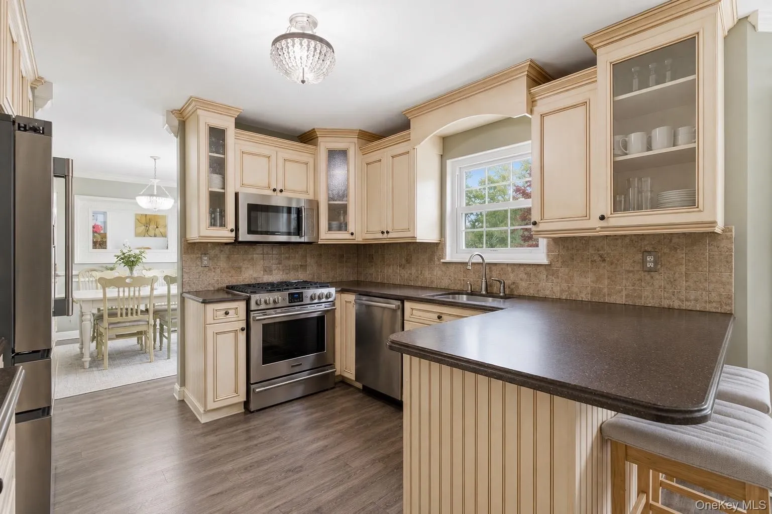 Kitchen featuring wood-finish flooring, stainless steel appliances, light-toned cabinetry with glass-front uppers, dark countertops, and a tiled backsplash Kitchen featuring wood-finish flooring, stainless steel appliances, light-toned cabinetry with glass-front uppers, dark countertops, and a tiled backsplash