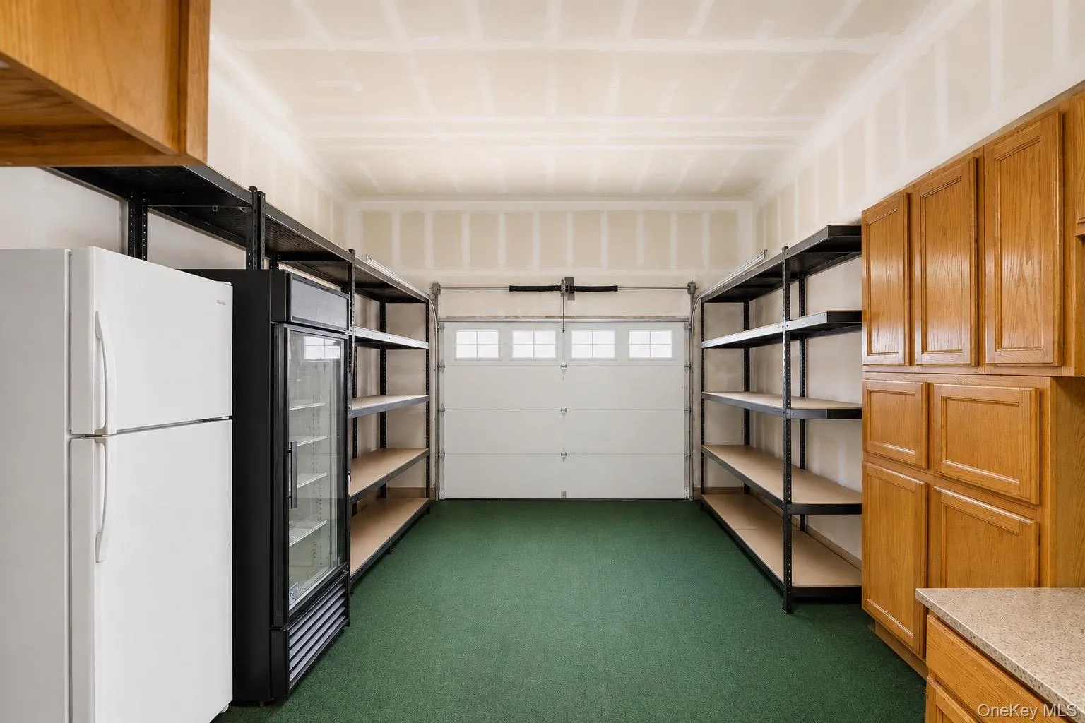 Garage space featuring a paneled overhead door, green carpet flooring, built-in wooden cabinetry, and extensive metal shelving systems Garage space featuring a paneled overhead door, green carpet flooring, built-in wooden cabinetry, and extensive metal shelving systems