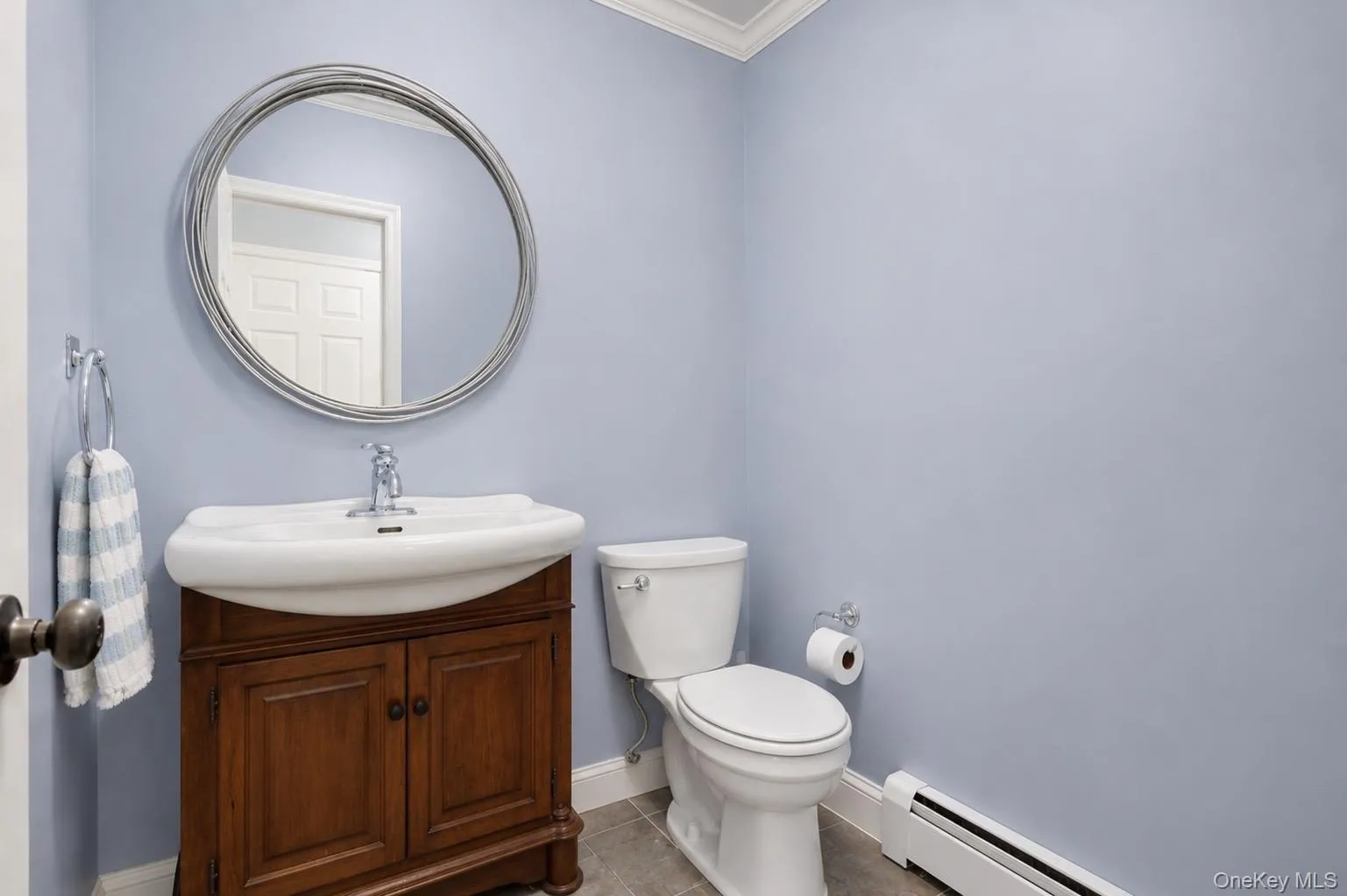 Bathroom featuring a wood-finish vanity with an integrated basin, a silver-framed circular mirror, a white toilet, and tile flooring Bathroom featuring a wood-finish vanity with an integrated basin, a silver-framed circular mirror, a white toilet, and tile flooring