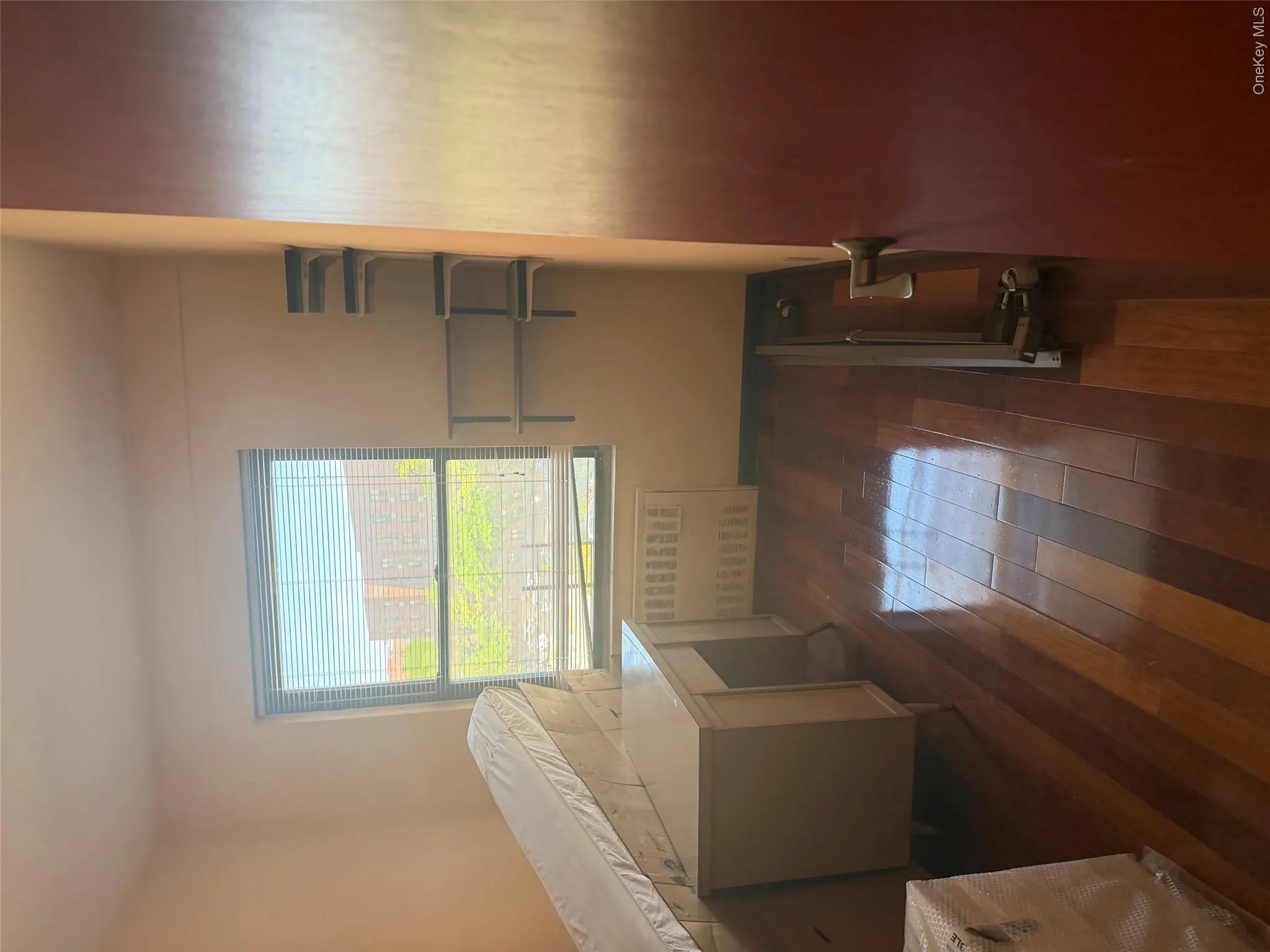 Wood-finish flooring throughout, featuring a single window with blinds, wall-mounted shelving, and a dark wood-finish interior door Wood-finish flooring throughout, featuring a single window with blinds, wall-mounted shelving, and a dark wood-finish interior door