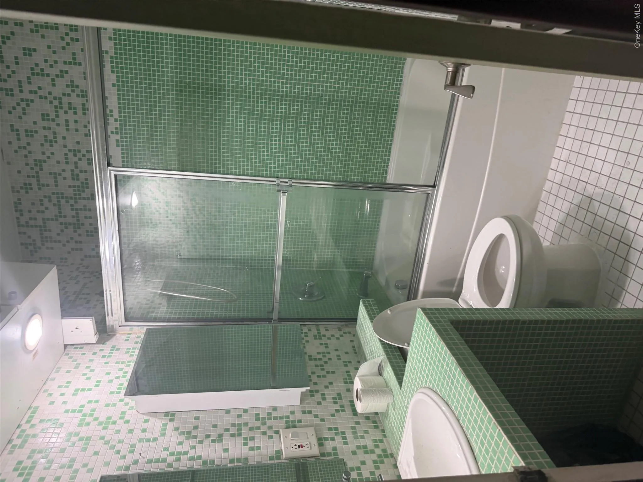 Bathroom with green and white mosaic tile walls, a white porcelain sink, a matching green tiled vanity, a white toilet, and a bathtub with a sliding glass door enclosure Bathroom with green and white mosaic tile walls, a white porcelain sink, a matching green tiled vanity, a white toilet, and a bathtub with a sliding glass door enclosure