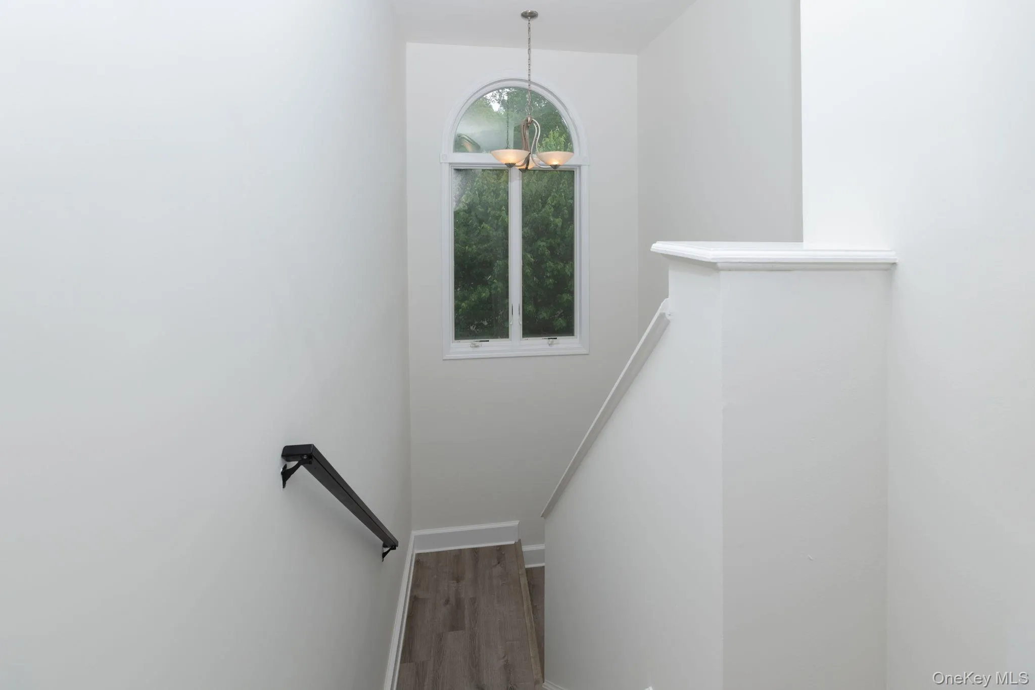 Stairwell featuring light wood-finish flooring, a black wall-mounted handrail, and a curved arch window with a chandelier Stairwell featuring light wood-finish flooring, a black wall-mounted handrail, and a curved arch window with a chandelier