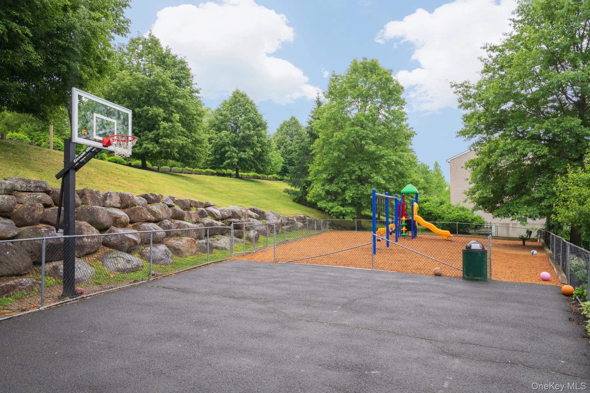 Paved recreation court featuring a basketball hoop, chain-link fencing, and a separate playground area with wood chip surfacing Paved recreation court featuring a basketball hoop, chain-link fencing, and a separate playground area with wood chip surfacing