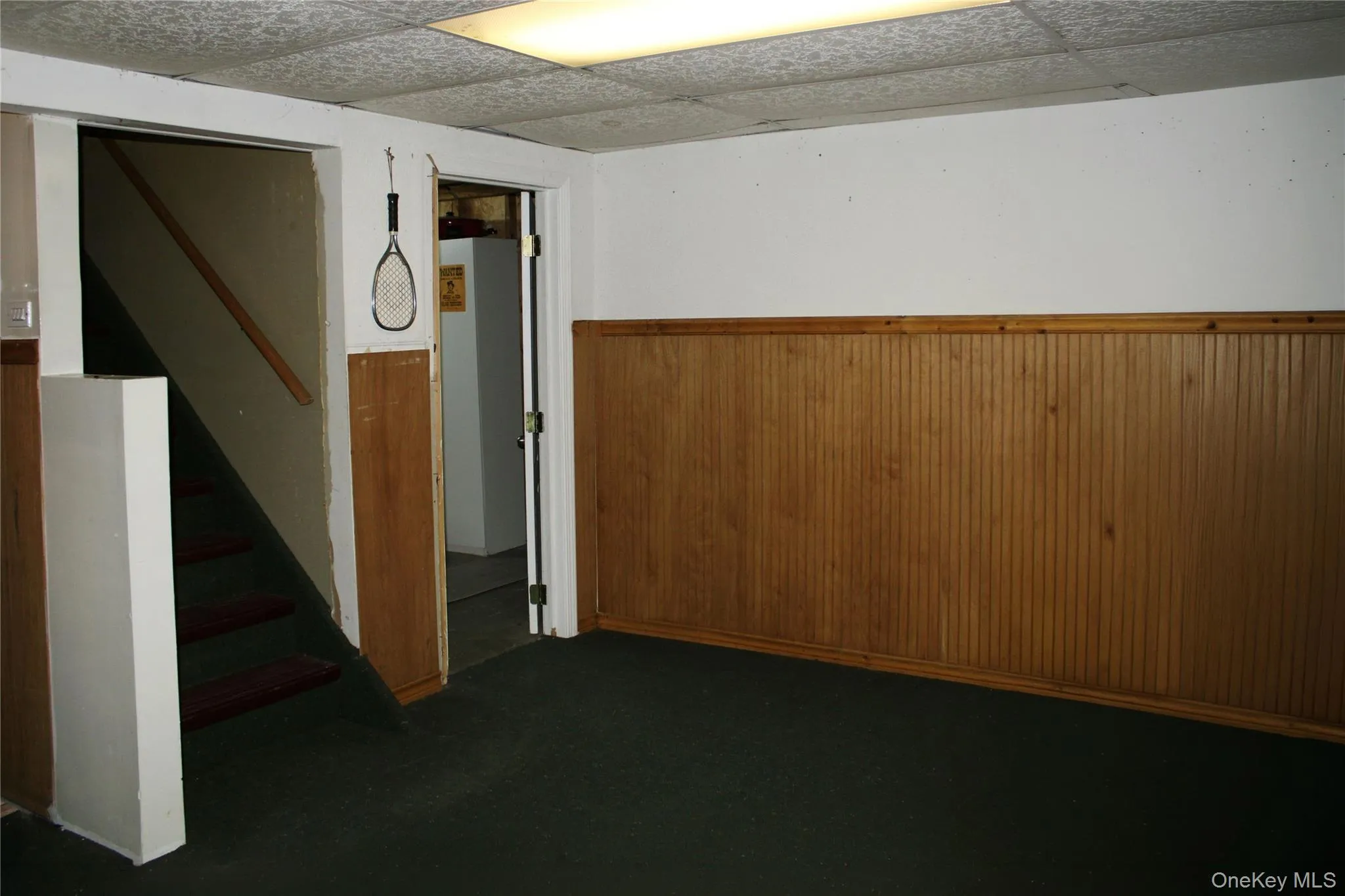 Recreational room featuring wood-finish beadboard paneling, white walls, dark green carpet flooring, and a drop ceiling with fluorescent lighting Recreational room featuring wood-finish beadboard paneling, white walls, dark green carpet flooring, and a drop ceiling with fluorescent lighting