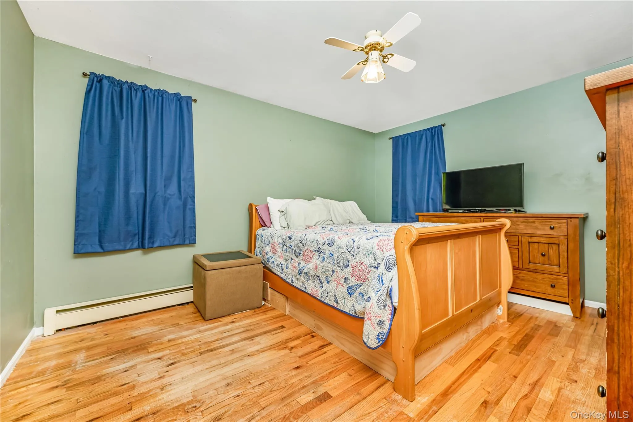 Room featuring wood-finish flooring, two windows with curtain rods, a ceiling fan with integrated lighting, and light green walls Room featuring wood-finish flooring, two windows with curtain rods, a ceiling fan with integrated lighting, and light green walls