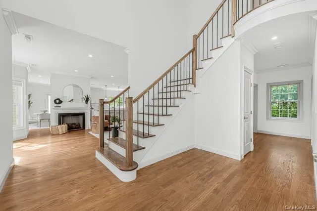 Grand foyer showcasing wood-finish flooring, a staircase with wood handrails and black metal balusters, crown molding, and a fireplace with a white mantel and black surround Grand foyer showcasing wood-finish flooring, a staircase with wood handrails and black metal balusters, crown molding, and a fireplace with a white mantel and black surround