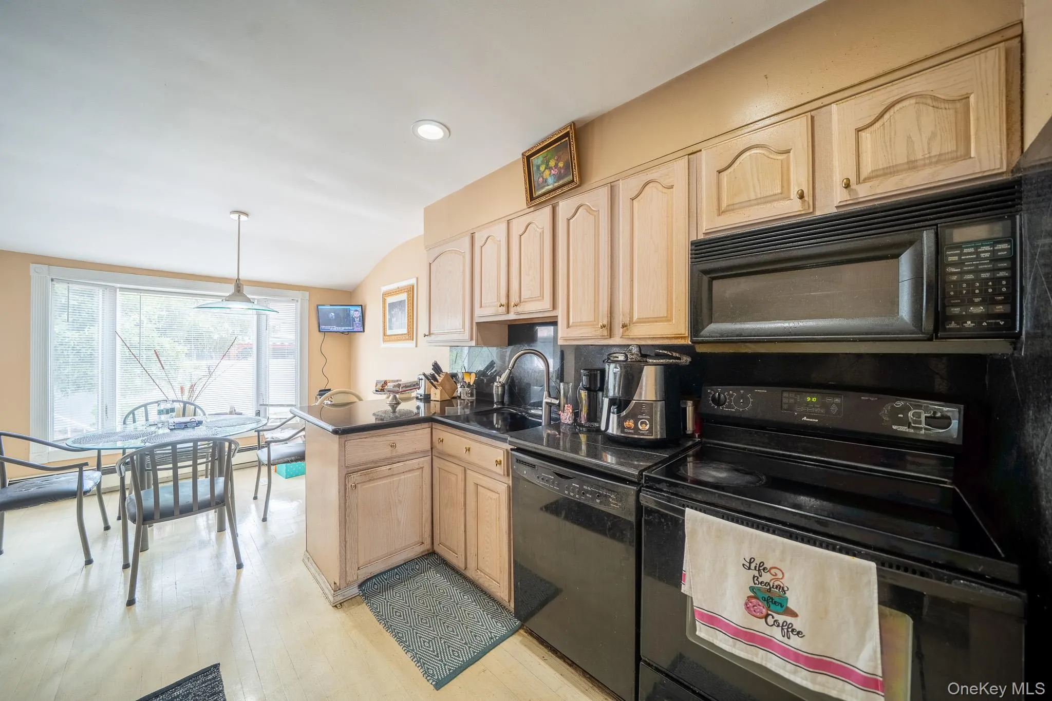 Kitchen with black appliances, light brown cabinets, a peninsula, dark countertops, and light wood finished floors Kitchen with black appliances, light brown cabinets, a peninsula, dark countertops, and light wood finished floors