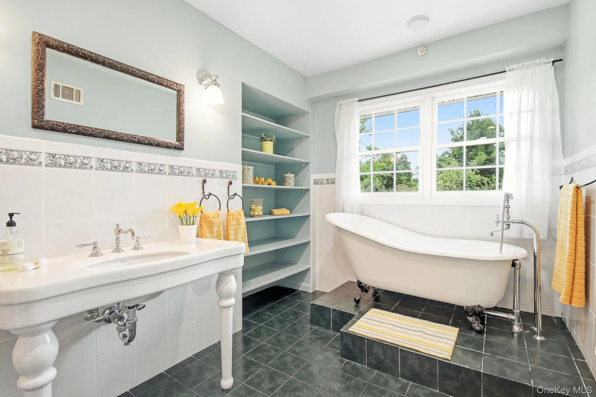 Primary en-suite bathroom featuring a freestanding clawfoot tub, a pedestal sink with chrome fixtures, built-in shelving, white tiled wainscoting with decorative trim, and dark tile flooring Primary en-suite bathroom featuring a freestanding clawfoot tub, a pedestal sink with chrome fixtures, built-in shelving, white tiled wainscoting with decorative trim, and dark tile flooring