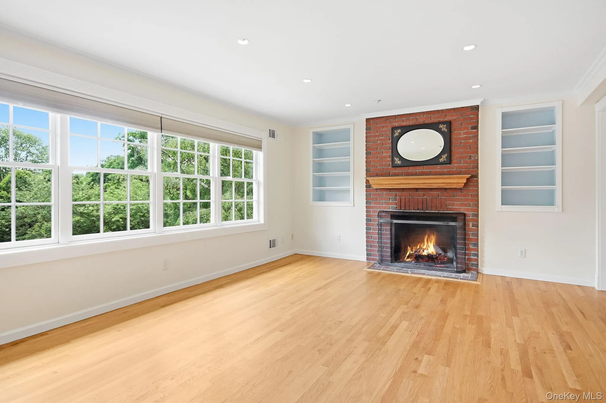 Spacious living room featuring hardwood flooring, a brick fireplace with a wood mantel, and built-in shelving Spacious living room featuring hardwood flooring, a brick fireplace with a wood mantel, and built-in shelving