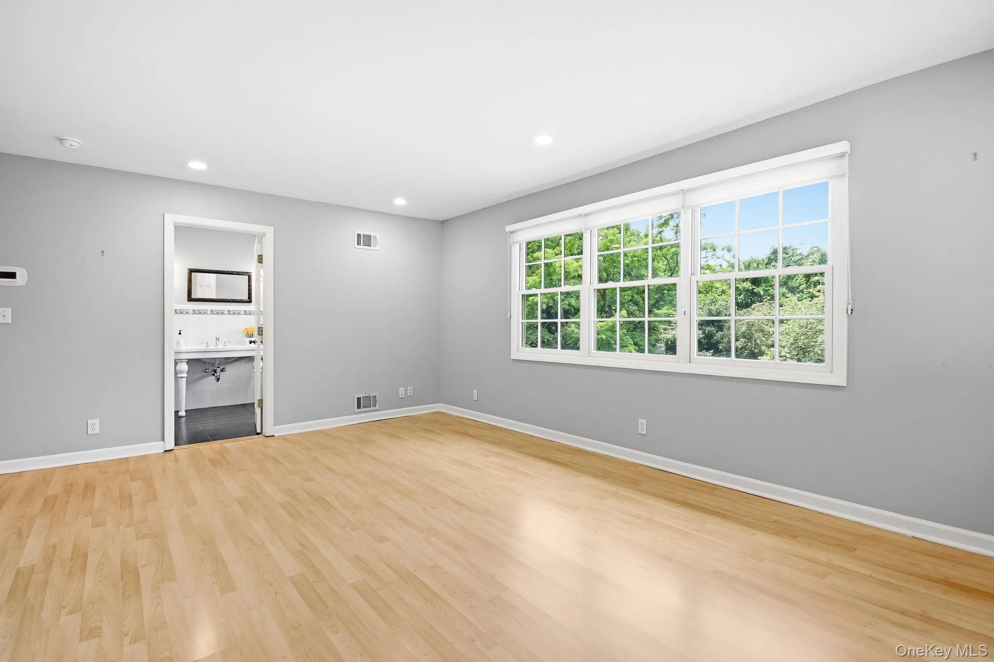 Primary en-suite (split level up); bright room featuring extensive natural light from multiple windows, recessed ceiling lighting, light wood-finish flooring, and light gray walls; alternate view towards primary bathroom. Primary en-suite (split level up); bright room featuring extensive natural light from multiple windows, recessed ceiling lighting, light wood-finish flooring, and light gray walls; alternate view towards primary bathroom.