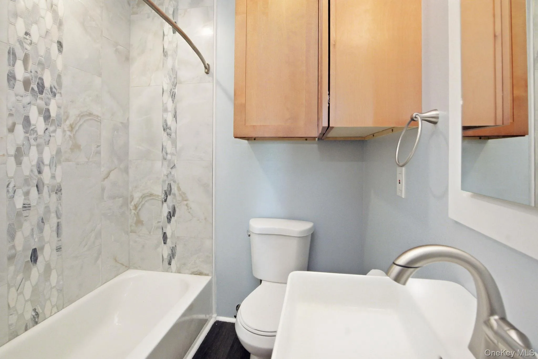 Bathroom featuring a white tub, shower with decorative hex tile accent, wood-finish flooring, wall-mounted cabinetry, and a modern vessel sink with a brushed nickel faucet. Everything in the apartment is completely brand new. Bathroom featuring a white tub, shower with decorative hex tile accent, wood-finish flooring, wall-mounted cabinetry, and a modern vessel sink with a brushed nickel faucet. Everything in the apartment is completely brand new.