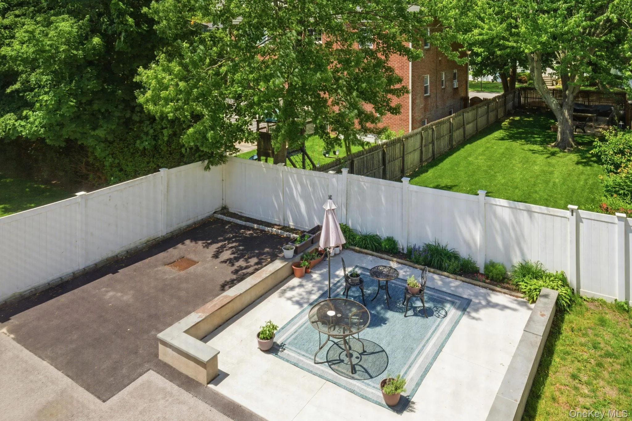 Expansive concrete patio area featuring a white privacy fence, an elevated planter, and surrounding green lawn Expansive concrete patio area featuring a white privacy fence, an elevated planter, and surrounding green lawn