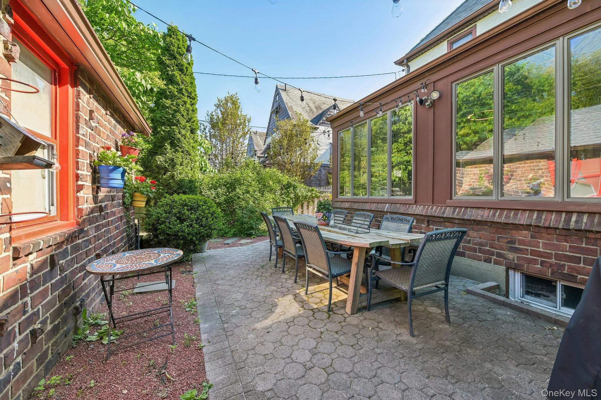 Paved patio area featuring a long dining table, surrounded by mature greenery Paved patio area featuring a long dining table, surrounded by mature greenery