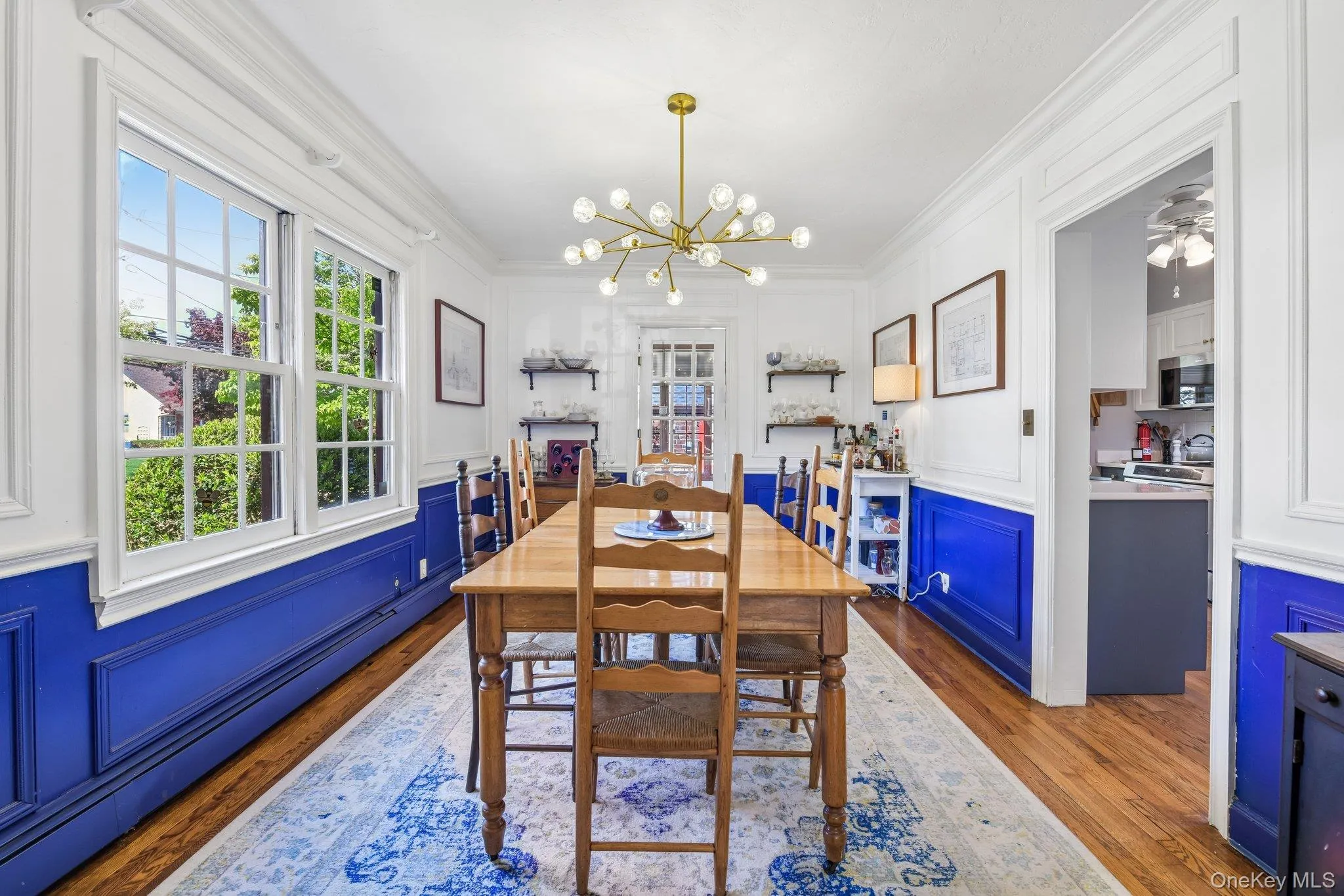 Open concept room featuring hardwood flooring, white paneled walls with blue wainscoting, a contemporary chandelier, and large window with exterior greenery Open concept room featuring hardwood flooring, white paneled walls with blue wainscoting, a contemporary chandelier, and large window with exterior greenery