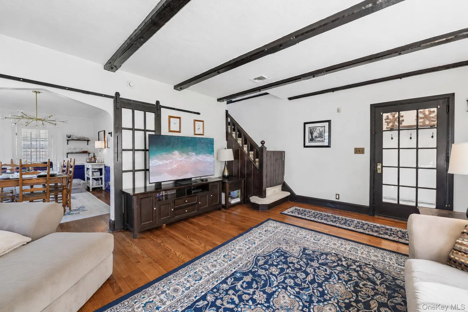 Living area featuring wood-finish flooring, exposed ceiling beams, and a staircase with wood newel posts Living area featuring wood-finish flooring, exposed ceiling beams, and a staircase with wood newel posts