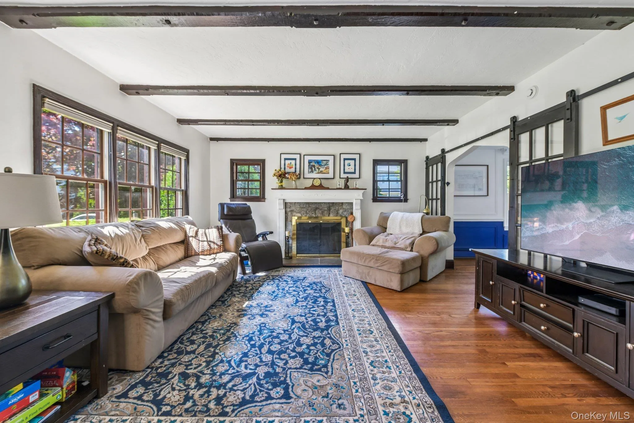 Living area featuring wood-finish flooring, exposed ceiling beams, a stone fireplace with a brass firebox, multiple windows with dark trim, and a built-in entertainment console Living area featuring wood-finish flooring, exposed ceiling beams, a stone fireplace with a brass firebox, multiple windows with dark trim, and a built-in entertainment console