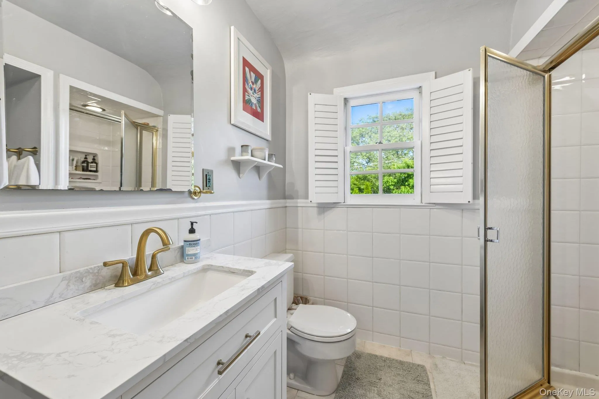 Bathroom with a white vanity featuring a rectangular sink and brass-finish faucet, a window with louvered shutters, a white toilet, and a glass-enclosed shower with brass-finish framing Bathroom with a white vanity featuring a rectangular sink and brass-finish faucet, a window with louvered shutters, a white toilet, and a glass-enclosed shower with brass-finish framing