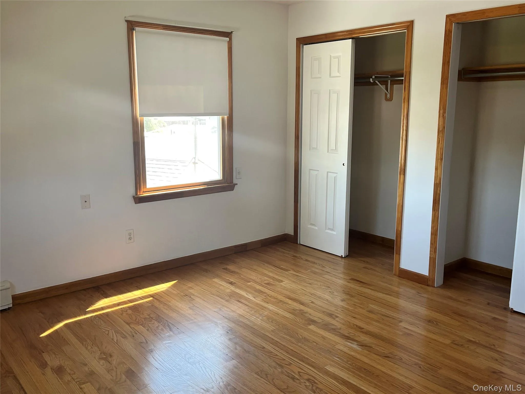 Room featuring wood-finish flooring, white walls, and a window with a roller shade Room featuring wood-finish flooring, white walls, and a window with a roller shade