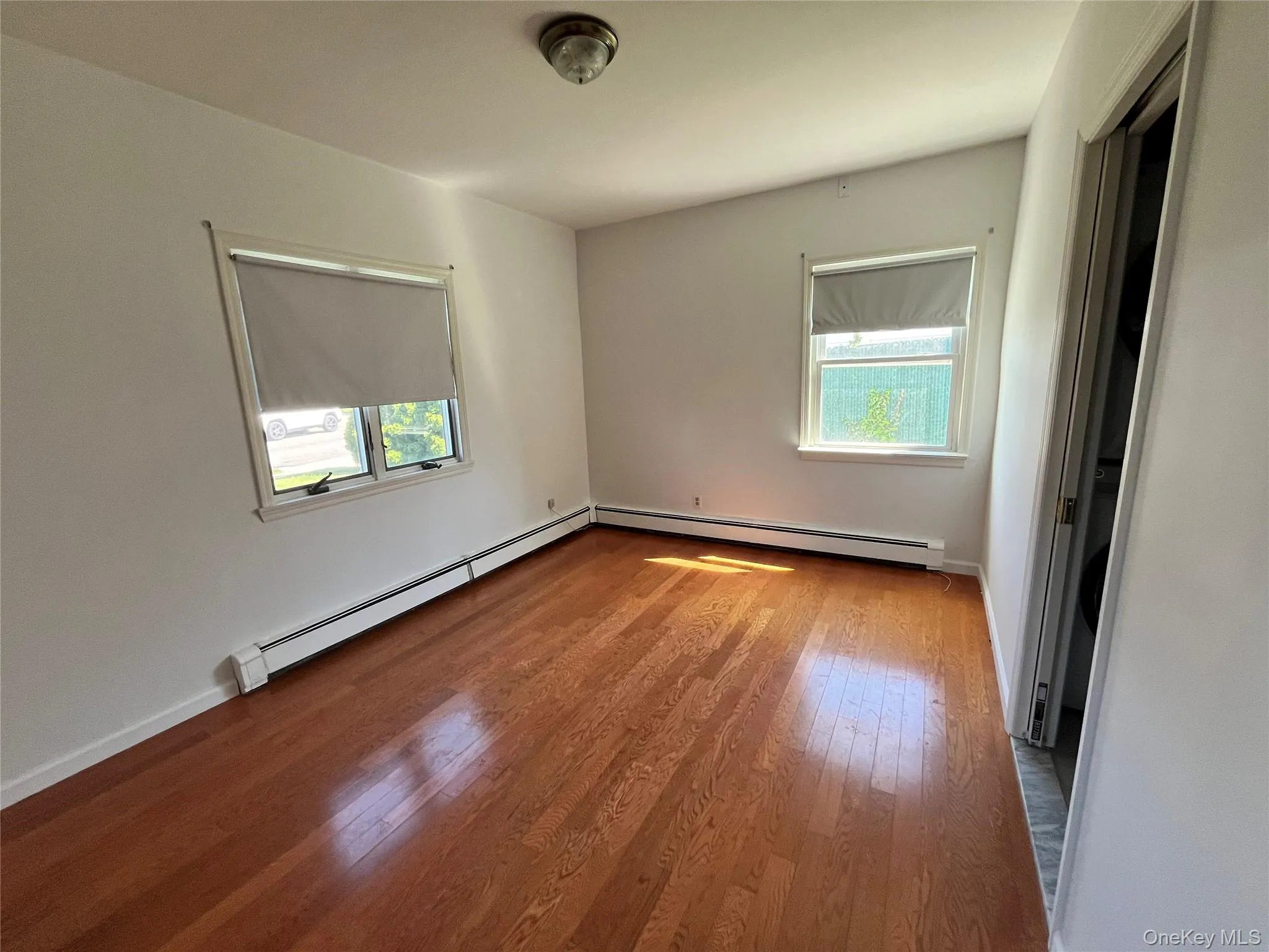 Hardwood flooring throughout, featuring two windows with roller shades, white baseboard trim, and a ceiling-mounted light fixture Hardwood flooring throughout, featuring two windows with roller shades, white baseboard trim, and a ceiling-mounted light fixture