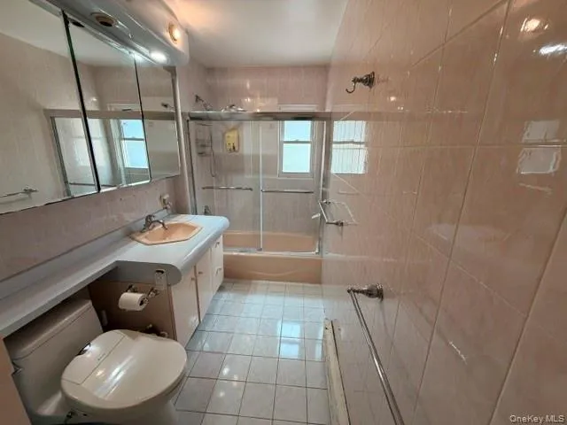 Full bathroom featuring a vanity with integrated sink, tri-fold mirrored medicine cabinet, a tiled shower/tub combination with sliding glass doors, and light-toned floor and wall tiling Full bathroom featuring a vanity with integrated sink, tri-fold mirrored medicine cabinet, a tiled shower/tub combination with sliding glass doors, and light-toned floor and wall tiling