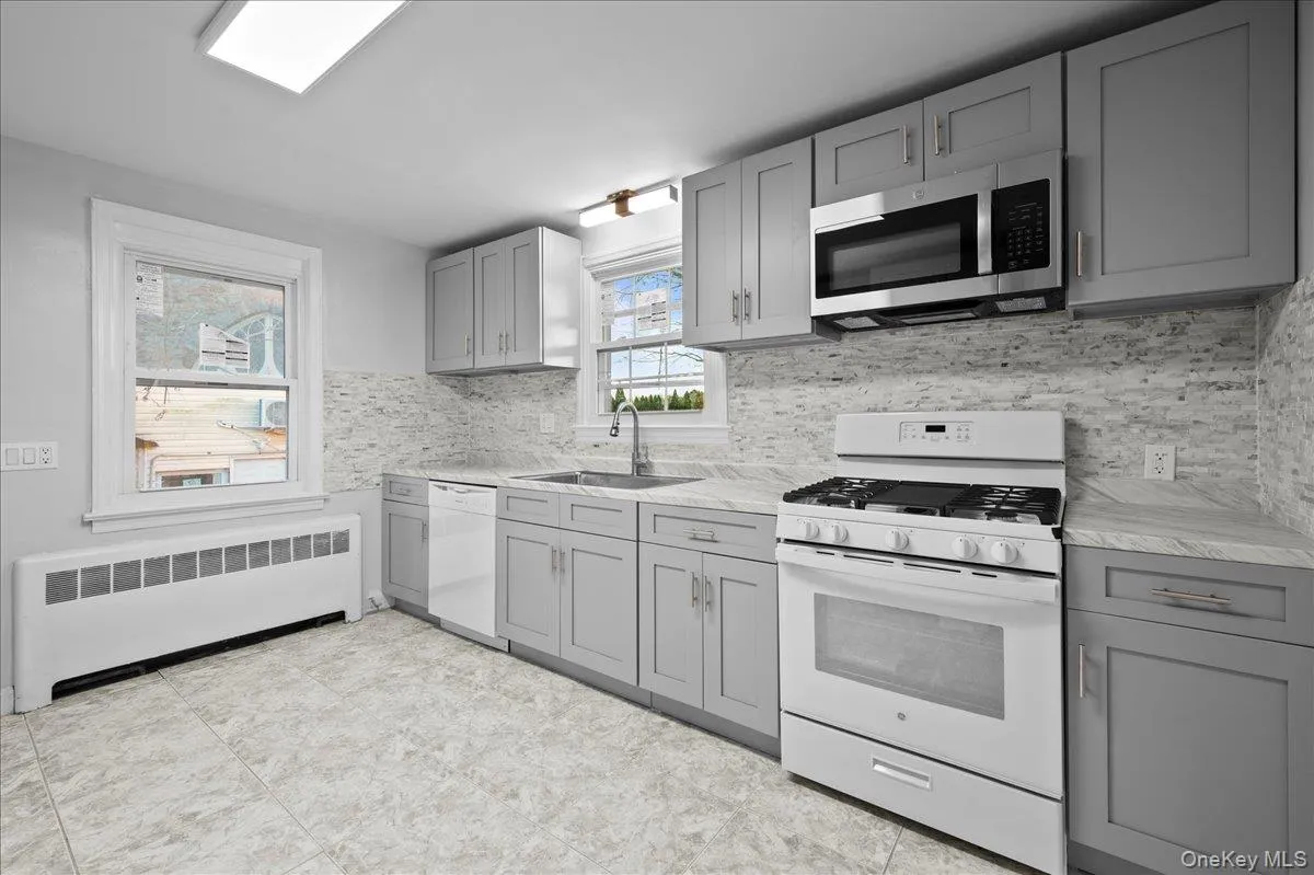 Kitchen featuring gray shaker-style cabinetry, white appliance suite, white tile backsplash, and light-toned countertops Kitchen featuring gray shaker-style cabinetry, white appliance suite, white tile backsplash, and light-toned countertops