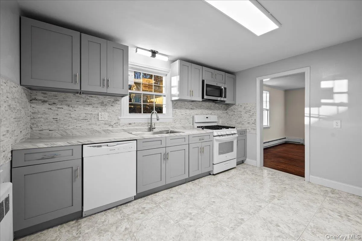 Kitchen featuring gray cabinetry, light-toned stone-look countertops, a tiled backsplash, and a white gas range with an overhead microwave Kitchen featuring gray cabinetry, light-toned stone-look countertops, a tiled backsplash, and a white gas range with an overhead microwave
