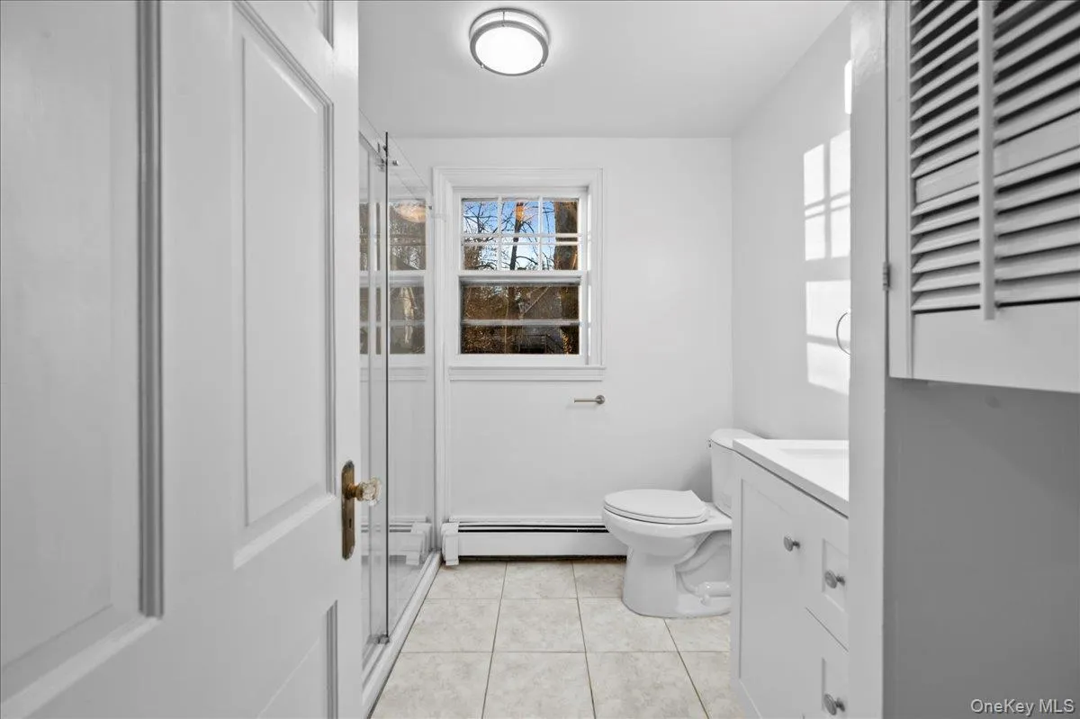 Bathroom featuring a shower enclosure with glass doors, a white vanity with an integrated sink, a louvered cabinet, a window, and tile flooring Bathroom featuring a shower enclosure with glass doors, a white vanity with an integrated sink, a louvered cabinet, a window, and tile flooring