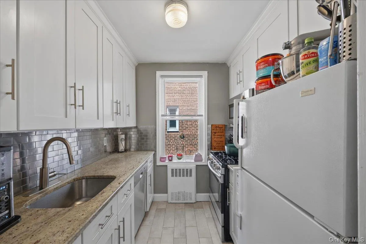 Galley kitchen featuring white cabinetry, a stainless steel sink with a gooseneck faucet, light-toned countertops, subway tile backsplash, and light wood-finish flooring Galley kitchen featuring white cabinetry, a stainless steel sink with a gooseneck faucet, light-toned countertops, subway tile backsplash, and light wood-finish flooring