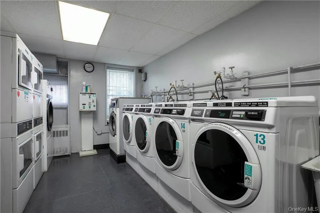 Communal laundry room featuring multiple front-loading washers and dryers, a drop ceiling with recessed lighting, and a window with security bars Communal laundry room featuring multiple front-loading washers and dryers, a drop ceiling with recessed lighting, and a window with security bars