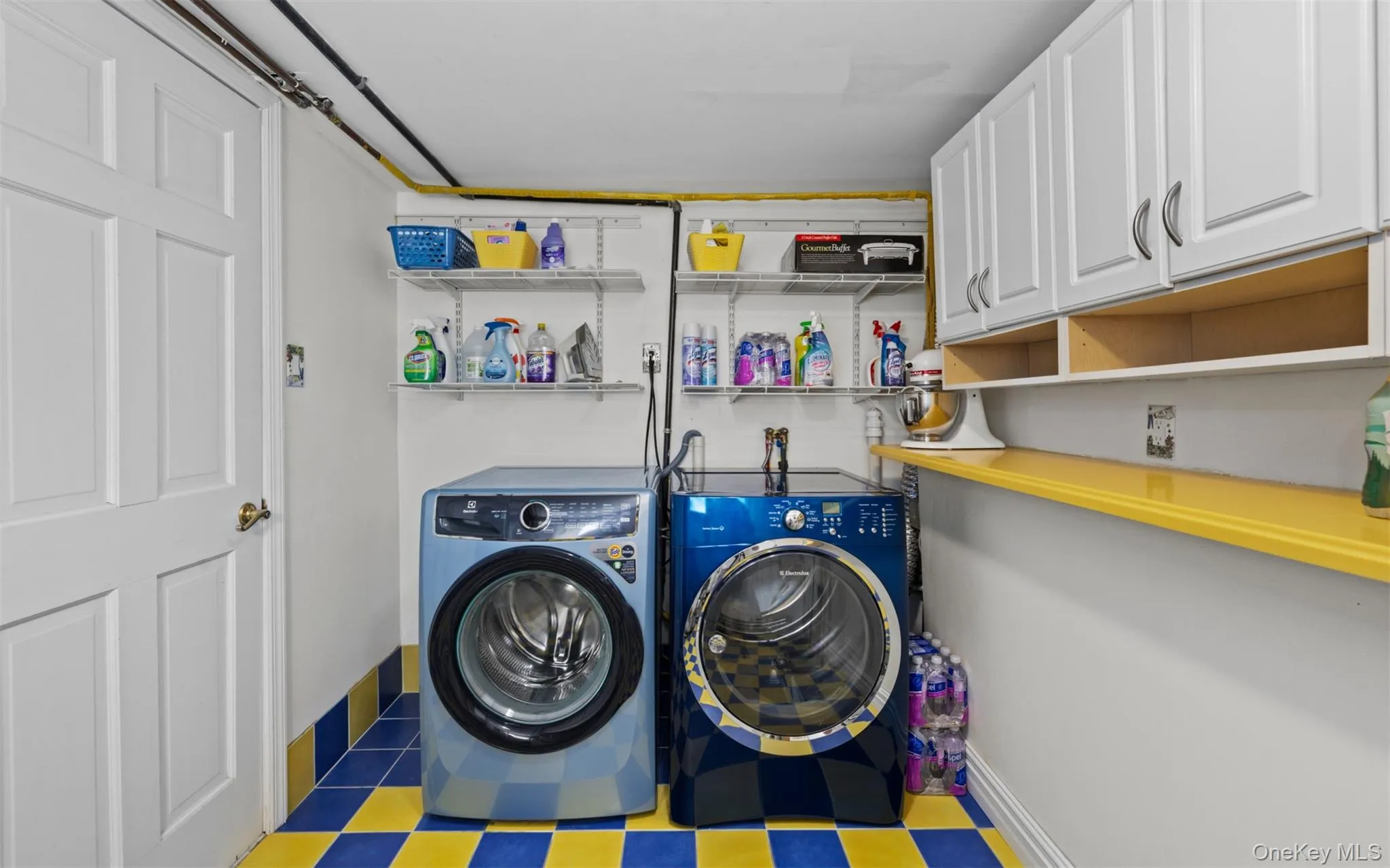 Laundry area featuring an overhead wire shelving system, wall-mounted white cabinetry, and a vibrant blue and yellow tiled floor Laundry area featuring an overhead wire shelving system, wall-mounted white cabinetry, and a vibrant blue and yellow tiled floor