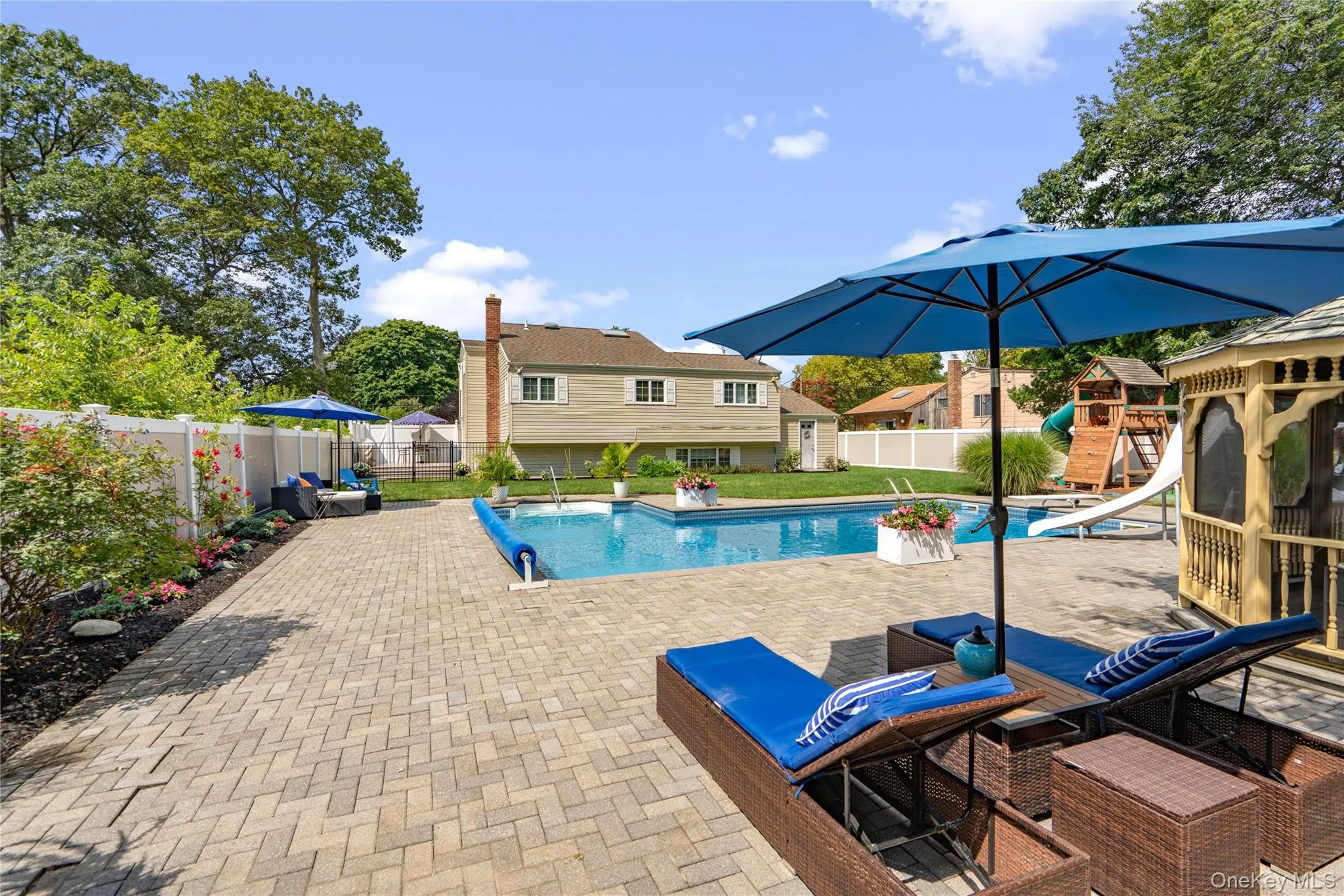 Expansive paver patio with an in-ground swimming pool, adjacent wooden gazebo, and a manicured planting bed bordering a white privacy fence Expansive paver patio with an in-ground swimming pool, adjacent wooden gazebo, and a manicured planting bed bordering a white privacy fence