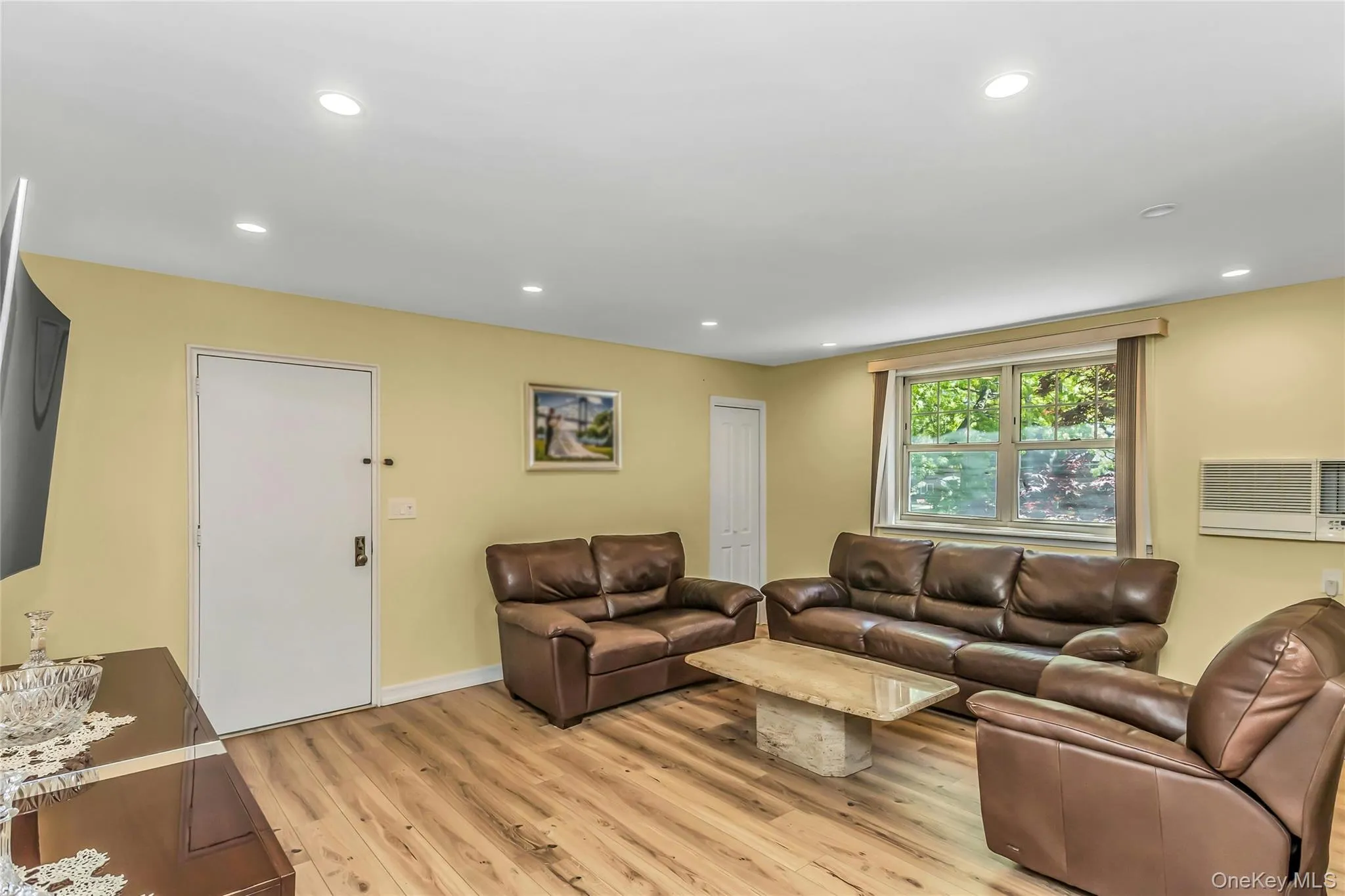 Spacious living area featuring wood-finish flooring, recessed ceiling lighting, a large window with natural light, and a built-in air conditioning unit Spacious living area featuring wood-finish flooring, recessed ceiling lighting, a large window with natural light, and a built-in air conditioning unit
