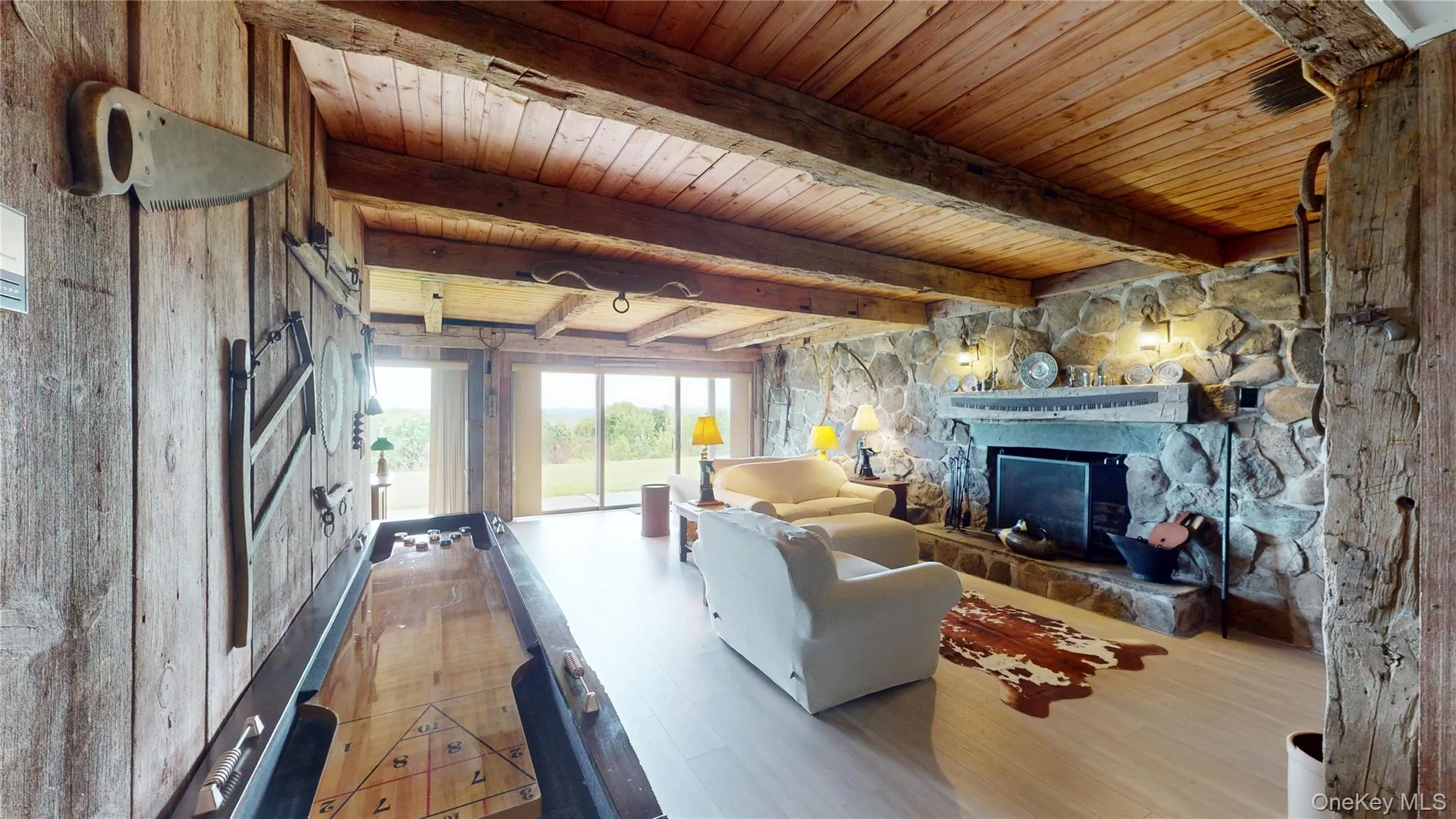 Spacious living area featuring a prominent stone fireplace, wood-planked walls, exposed wood beams, wood-finish flooring, and expansive glass sliding doors Spacious living area featuring a prominent stone fireplace, wood-planked walls, exposed wood beams, wood-finish flooring, and expansive glass sliding doors
