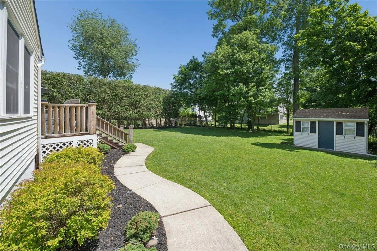 Expansive rear yard featuring a wooden deck with railing and stairs, a winding concrete pathway, mature perimeter hedging, and a separate storage shed with a shingled roof. Expansive rear yard featuring a wooden deck with railing and stairs, a winding concrete pathway, mature perimeter hedging, and a separate storage shed with a shingled roof.