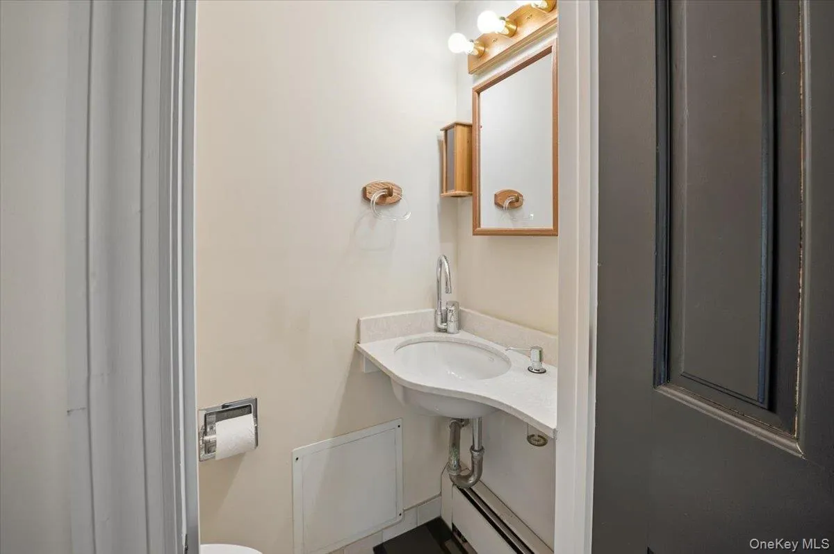 Compact bathroom featuring a corner-mounted sink with a light-toned countertop, a wall-mounted mirror, and overhead vanity lighting Compact bathroom featuring a corner-mounted sink with a light-toned countertop, a wall-mounted mirror, and overhead vanity lighting