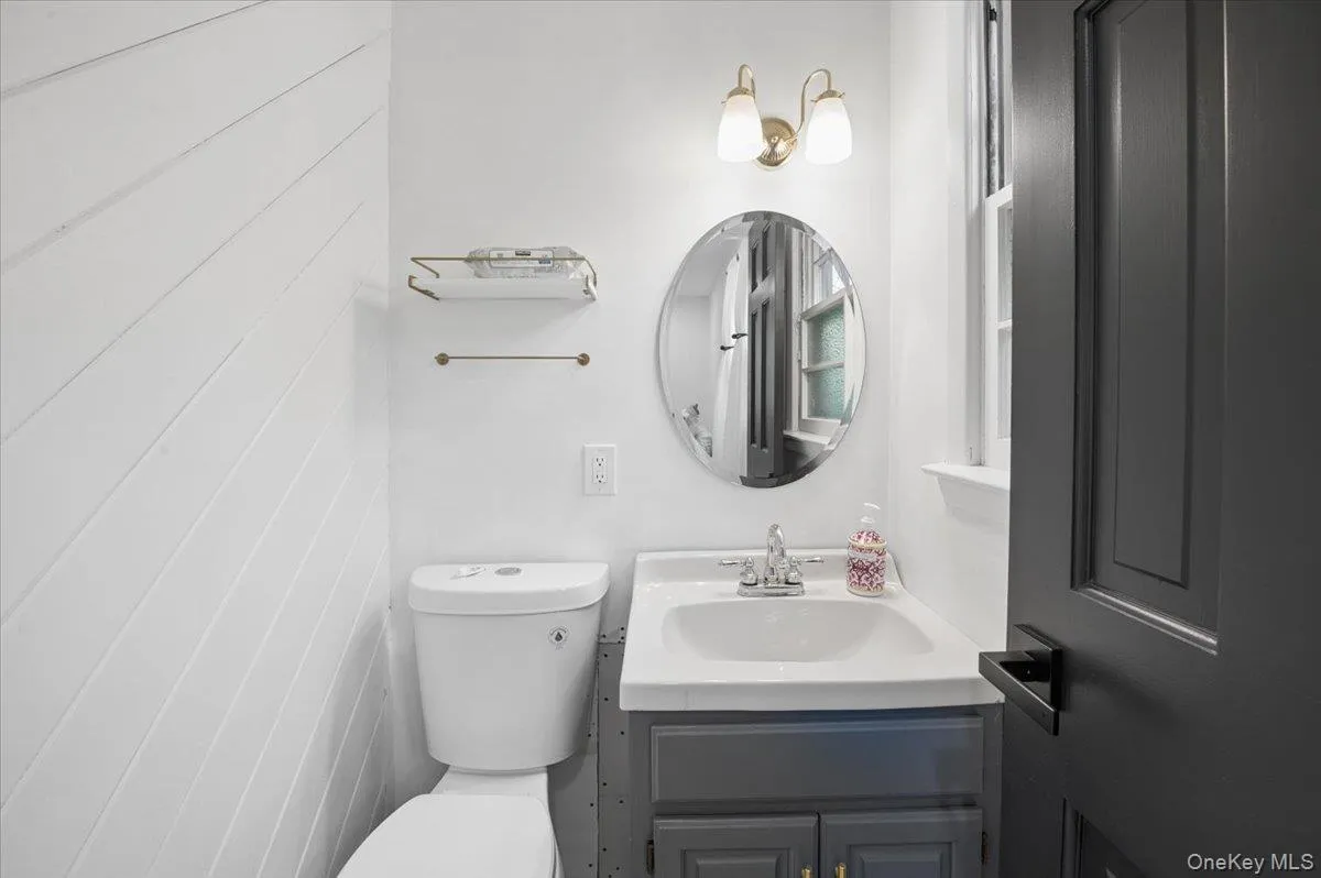 Bathroom featuring a white toilet, gray vanity with integrated sink, oval mirror, and brass vanity light. Bathroom featuring a white toilet, gray vanity with integrated sink, oval mirror, and brass vanity light.