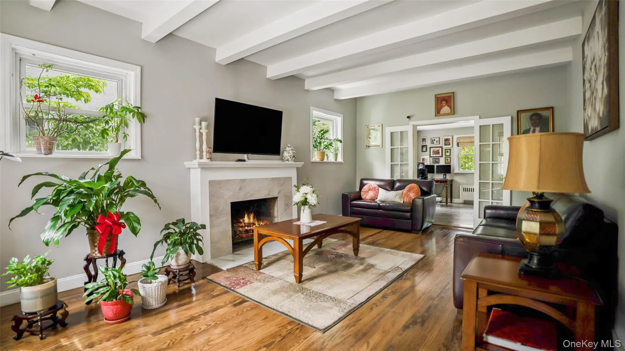Living room featuring wood-finish flooring, a fireplace with a stone surround and white mantle, exposed ceiling beams, and dual windows Living room featuring wood-finish flooring, a fireplace with a stone surround and white mantle, exposed ceiling beams, and dual windows