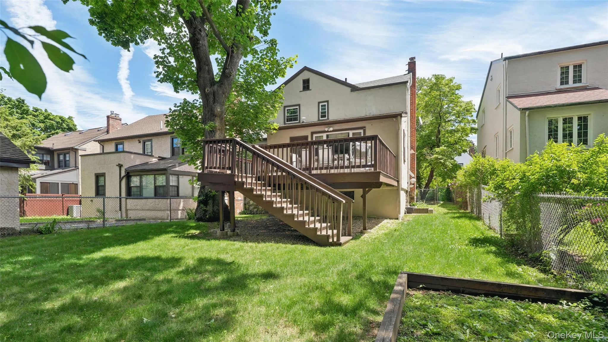 Expansive backyard featuring a tiered wooden deck with railings and stairs, a large mature tree, manicured lawn, and perimeter chain link fencing Expansive backyard featuring a tiered wooden deck with railings and stairs, a large mature tree, manicured lawn, and perimeter chain link fencing