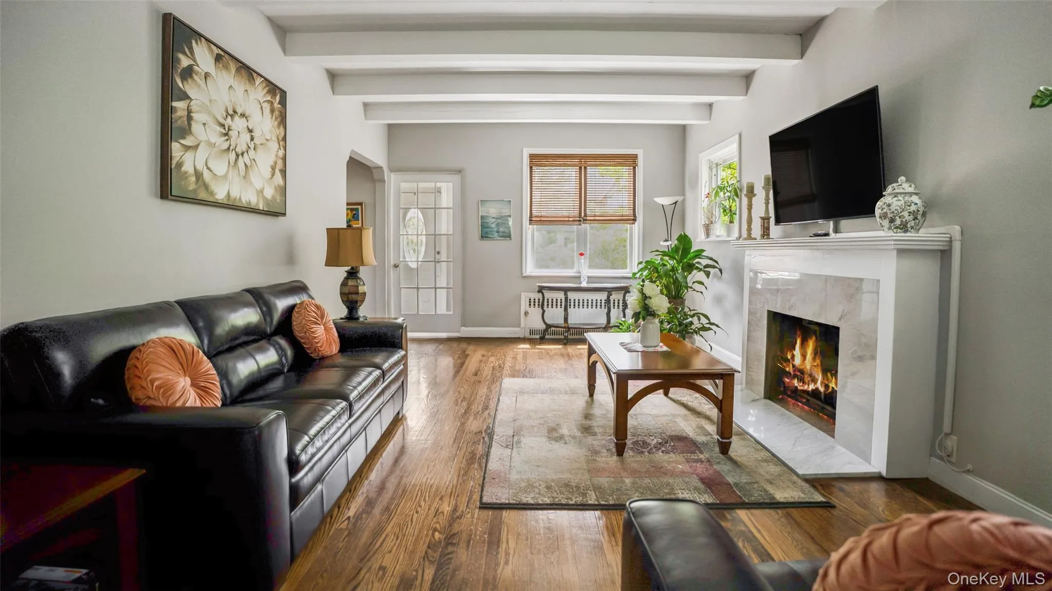 Living room featuring a fireplace with a stone surround, wood-finish flooring, exposed ceiling beams, and light-toned walls Living room featuring a fireplace with a stone surround, wood-finish flooring, exposed ceiling beams, and light-toned walls