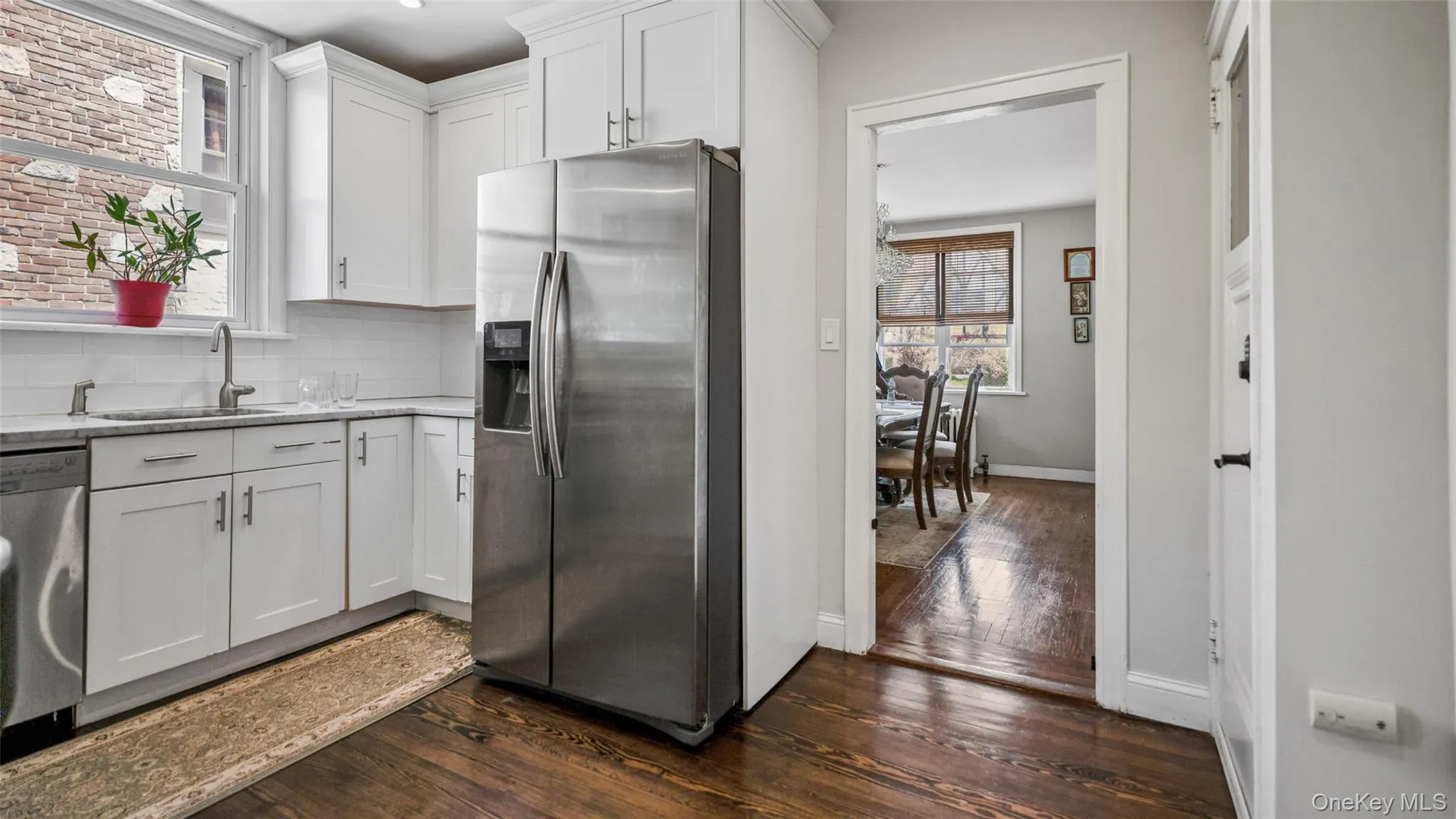 Kitchen featuring white shaker cabinetry, stainless steel refrigerator, undermount sink with gooseneck faucet, light-toned countertop, and dark wood-finish flooring Kitchen featuring white shaker cabinetry, stainless steel refrigerator, undermount sink with gooseneck faucet, light-toned countertop, and dark wood-finish flooring