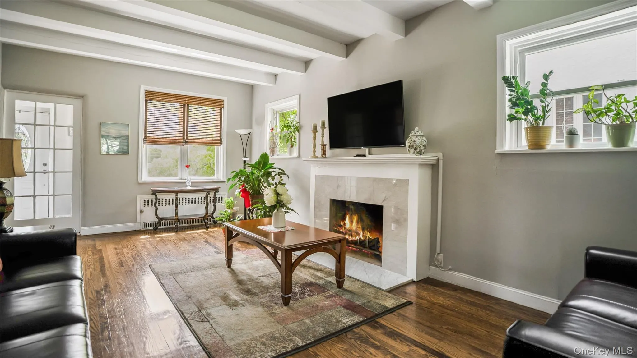 Spacious living area featuring exposed ceiling beams, wood-finish flooring, and a fireplace with a stone surround and white mantel Spacious living area featuring exposed ceiling beams, wood-finish flooring, and a fireplace with a stone surround and white mantel
