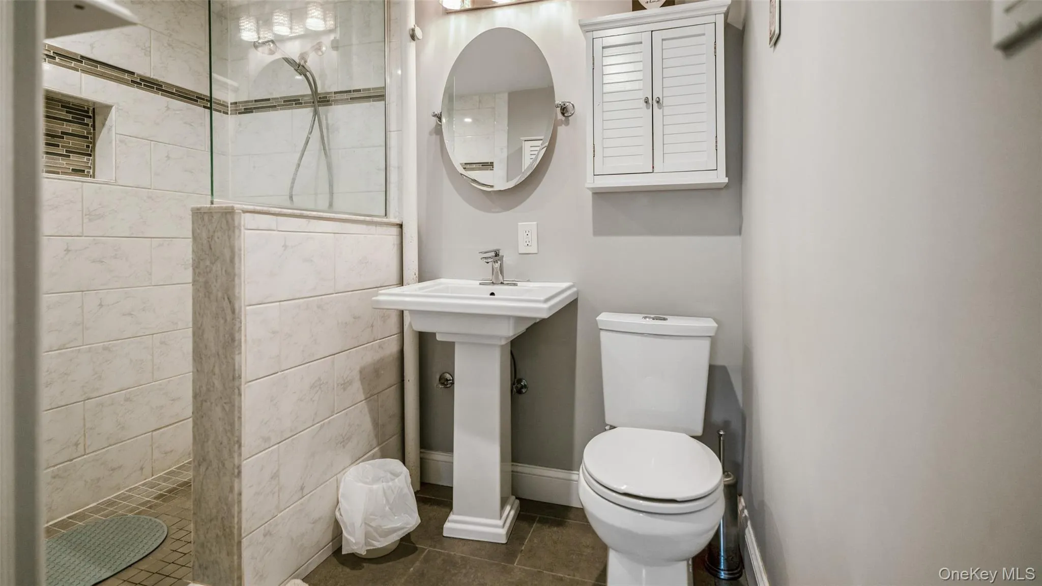 Bathroom featuring a glass-enclosed shower with tiled walls and a recessed niche, a white pedestal sink, an oval wall-mounted mirror, and a white louvered wall cabinet Bathroom featuring a glass-enclosed shower with tiled walls and a recessed niche, a white pedestal sink, an oval wall-mounted mirror, and a white louvered wall cabinet